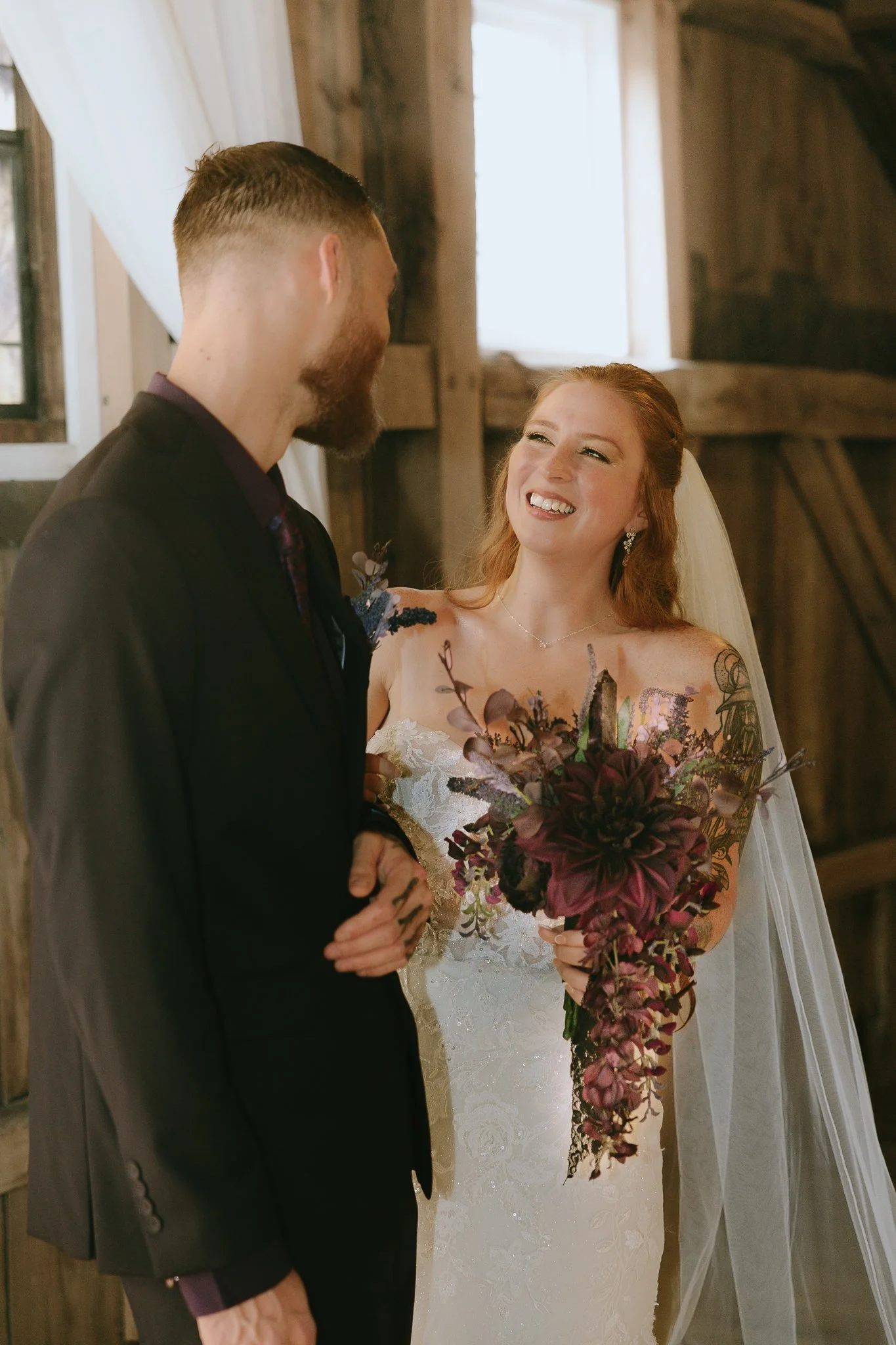 Bride and groom facing each other indoors, smiling, with the bride holding a large bouquet of dark purple and pink flowers.