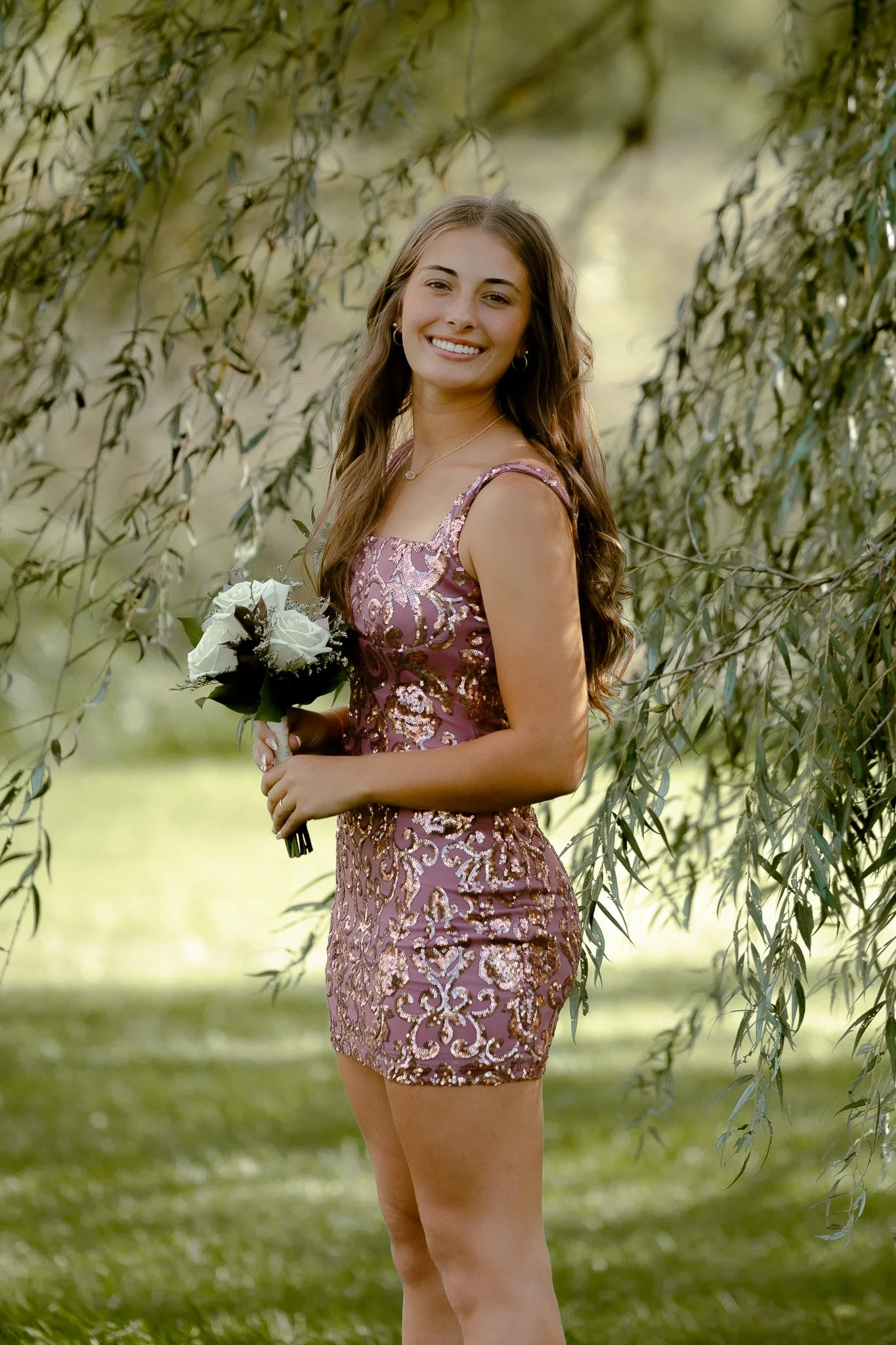 A young woman with long brown hair wearing a pink and gold sequined dress, smiling and holding a bouquet of white roses, standing outdoors near a tree with green leaves.