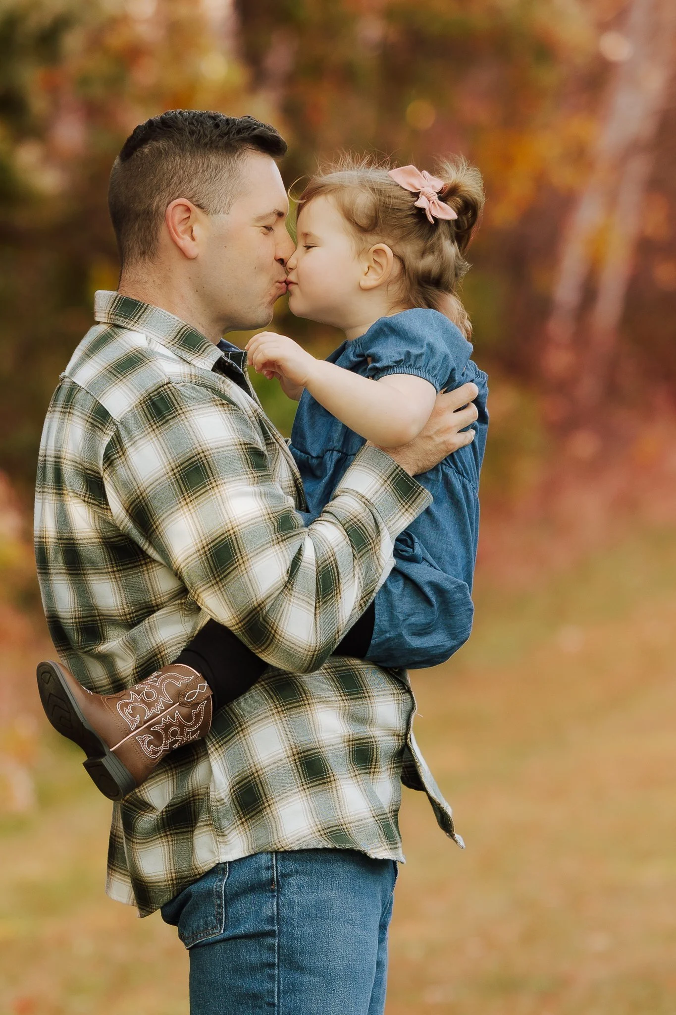 A man holding a young girl in his arms as they share a kiss outdoors during fall, with colorful autumn foliage in the background.