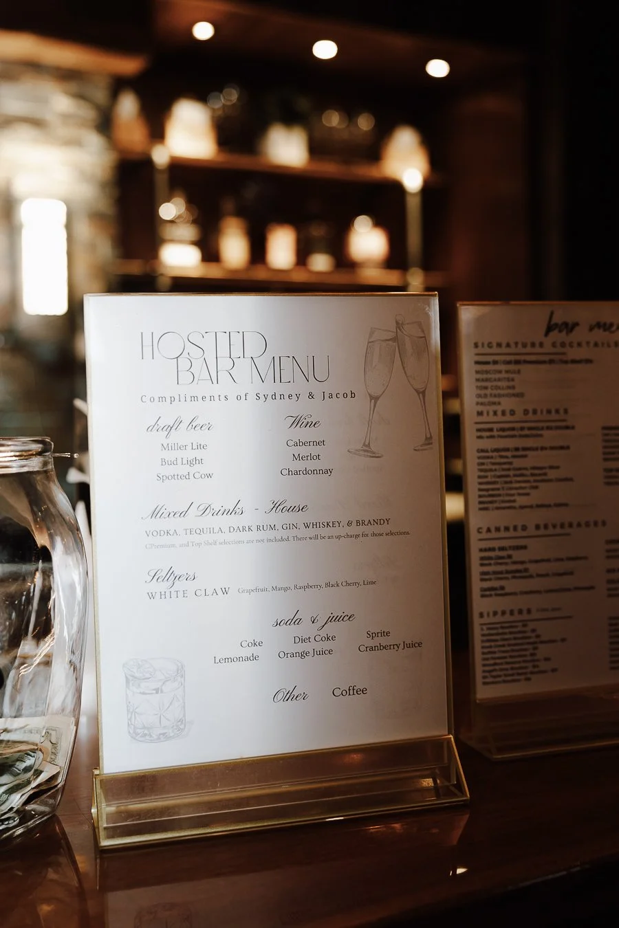 Restaurant bar menu on a stand, listing wine, mixed drinks, seltzers, soda, and coffee, with a jar of water and a wooden table.