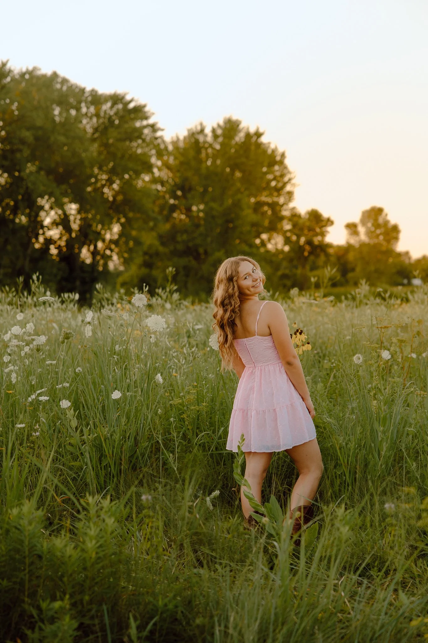 A woman in a pink dress standing in a grassy field with wildflowers, smiling and looking back at the camera at sunset.
