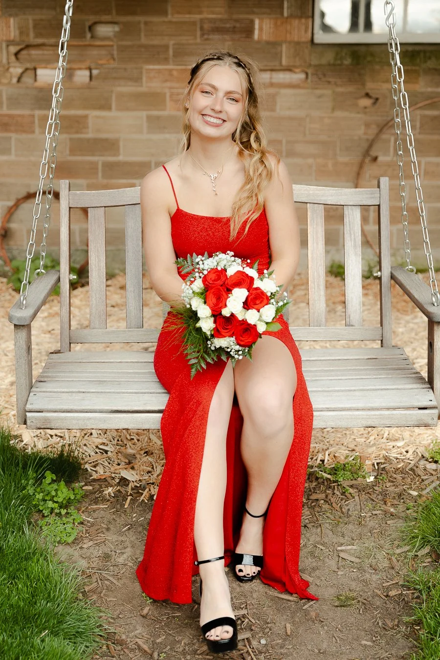 A young woman in a red dress sitting on a wooden swing, holding a bouquet of red and white roses, smiling at the camera.