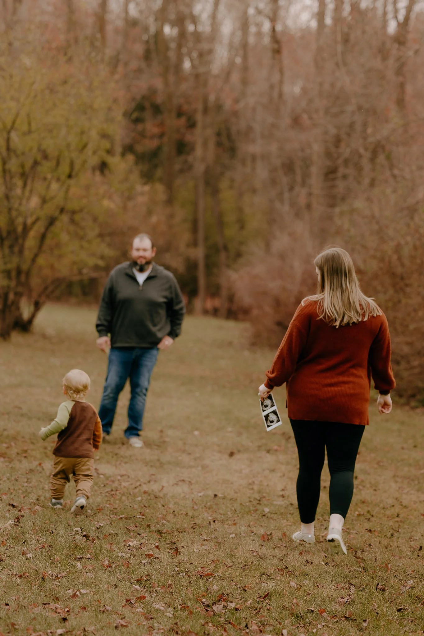 A family walking outdoors in an autumn setting, with trees with orange leaves in the background. The woman is holding an ultrasound picture, and the child is running toward the man.