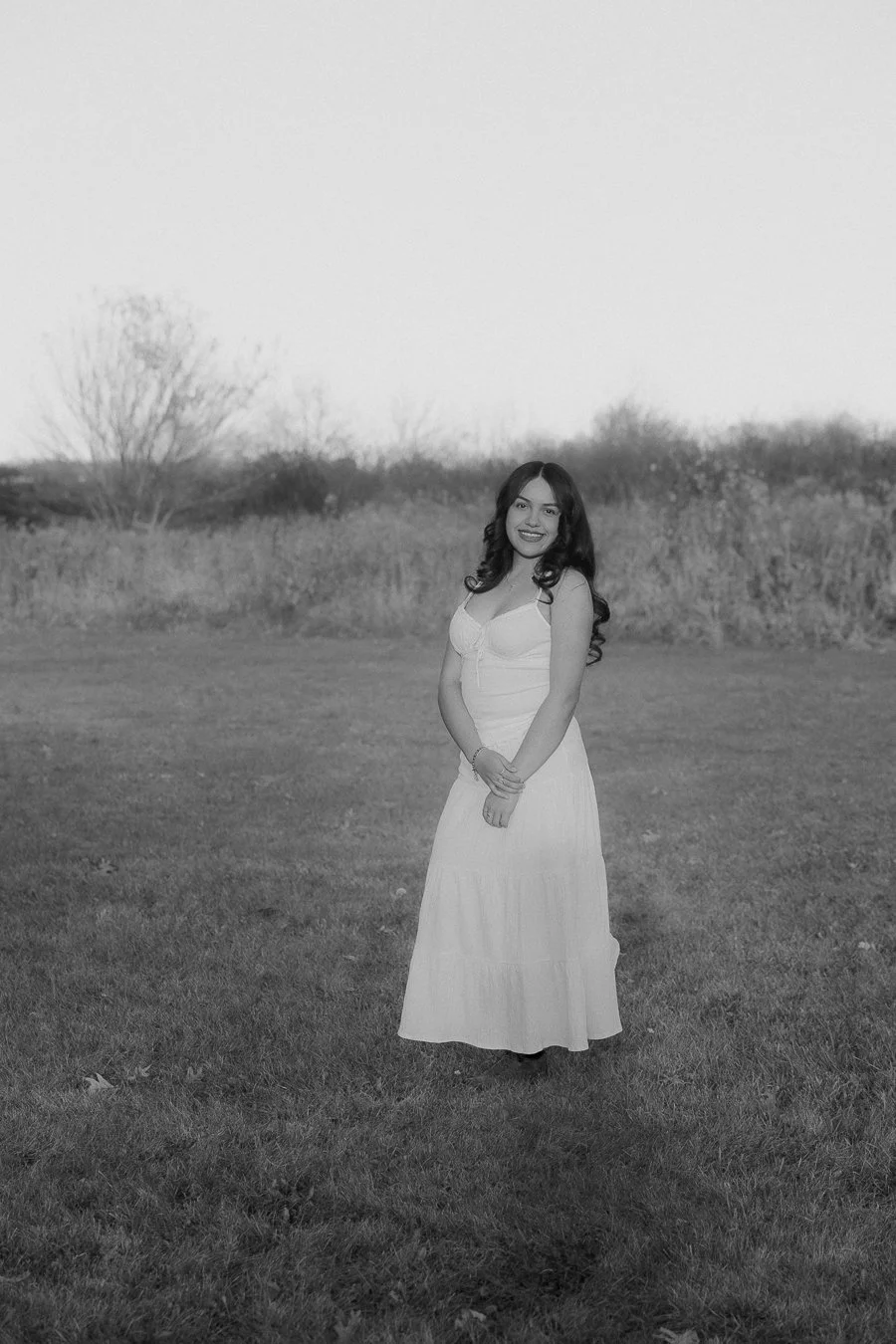 A young woman in a white dress standing outdoors on a grassy field, smiling at the camera, with trees and bushes in the background.