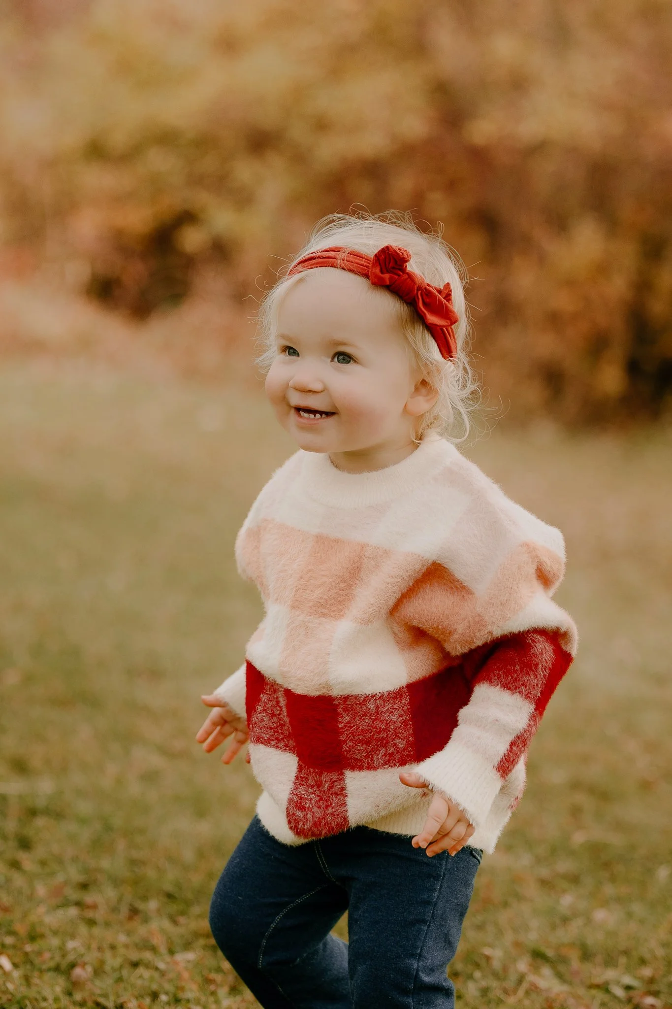 A young girl with curly blonde hair wearing a red headband with a bow, a cozy cream and red plaid sweater, and dark jeans, smiling outdoors on a fall day with autumn leaves in the background.