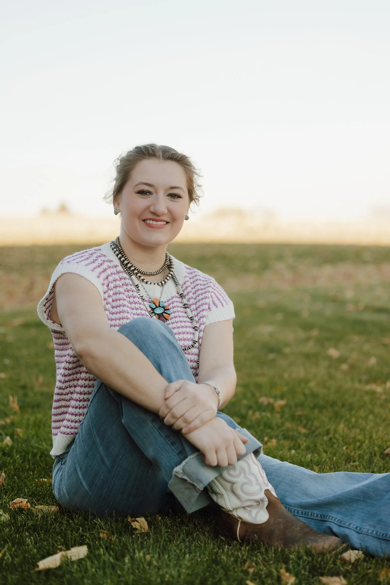 A young woman sitting on grass outdoors, smiling at the camera, wearing a striped pink and white top, jeans, cowboy boots, and layered jewelry, with a clear sky and trees in the background.