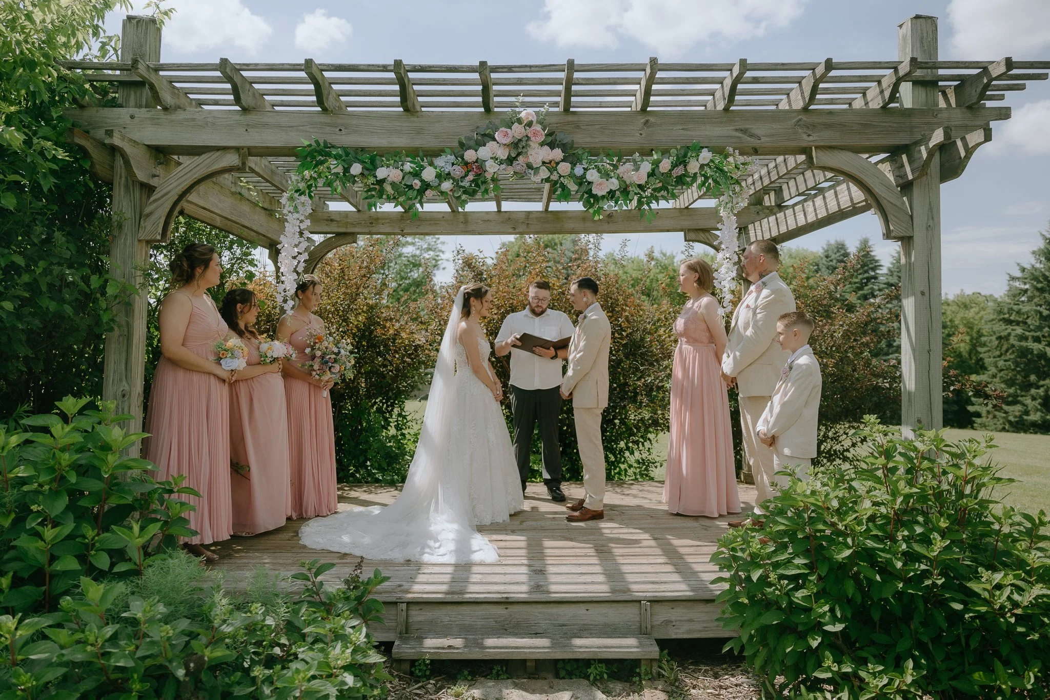 Outdoor wedding ceremony with a bride and groom exchanging vows under a wooden arbor decorated with flowers, surrounded by bridesmaids and groomsmen, on a sunny day with green trees in the background.