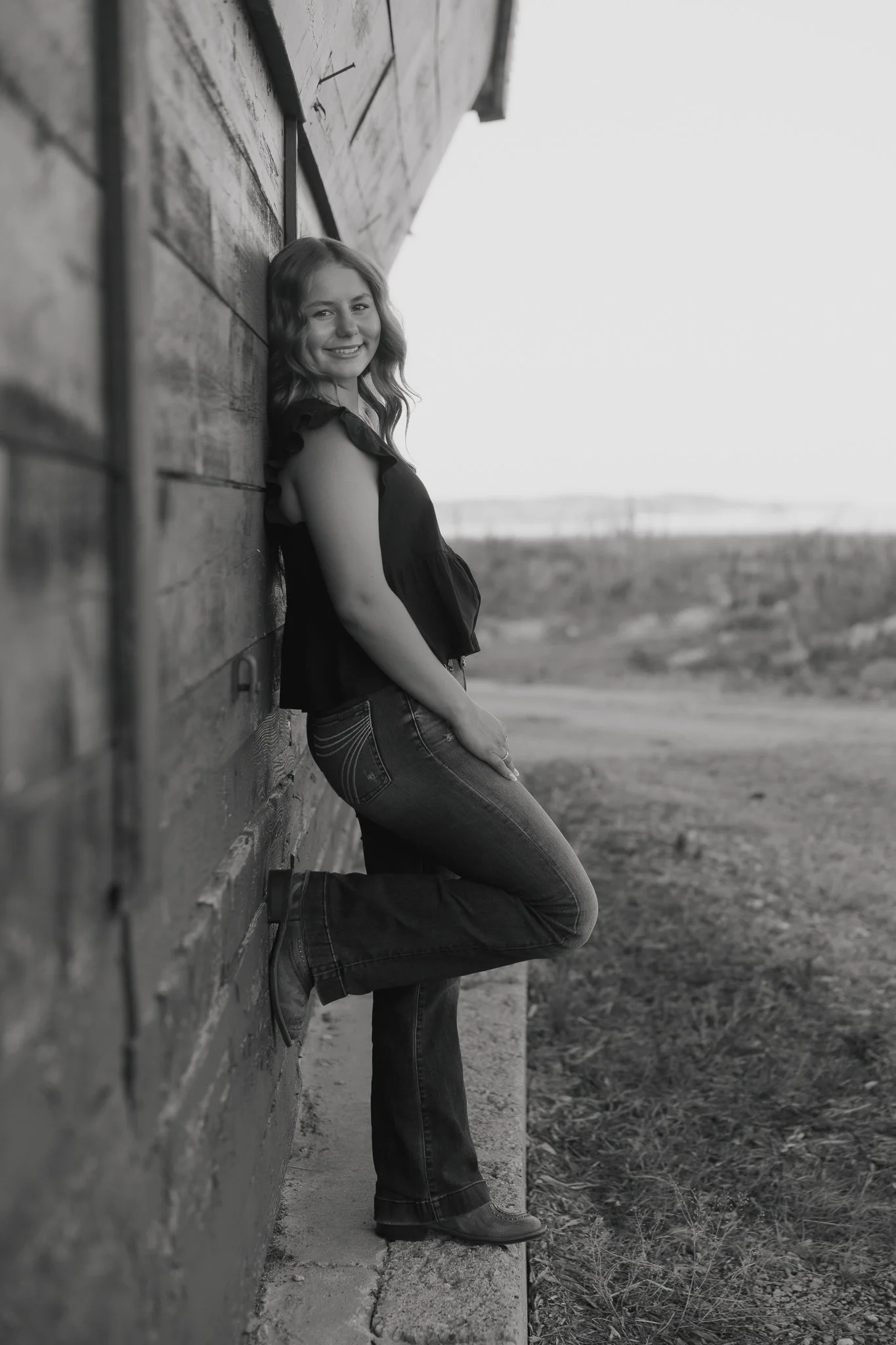 Black and white photo of a smiling young woman leaning against a wooden wall outdoors, with open fields in the background.