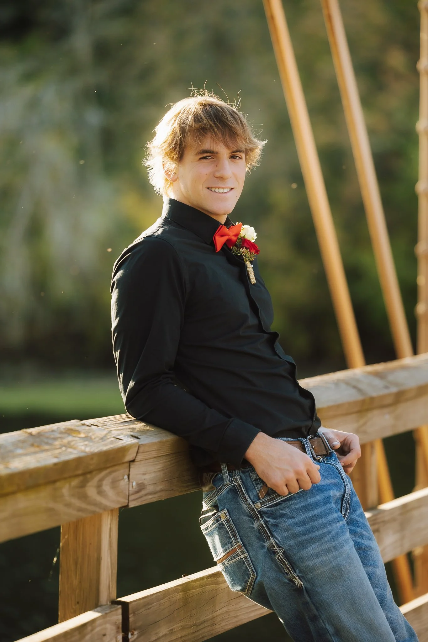 A young man in a black shirt and blue jeans leaning against a wooden railing outdoors at sunset, wearing a red bow tie and floral boutonniere, smiling at the camera.