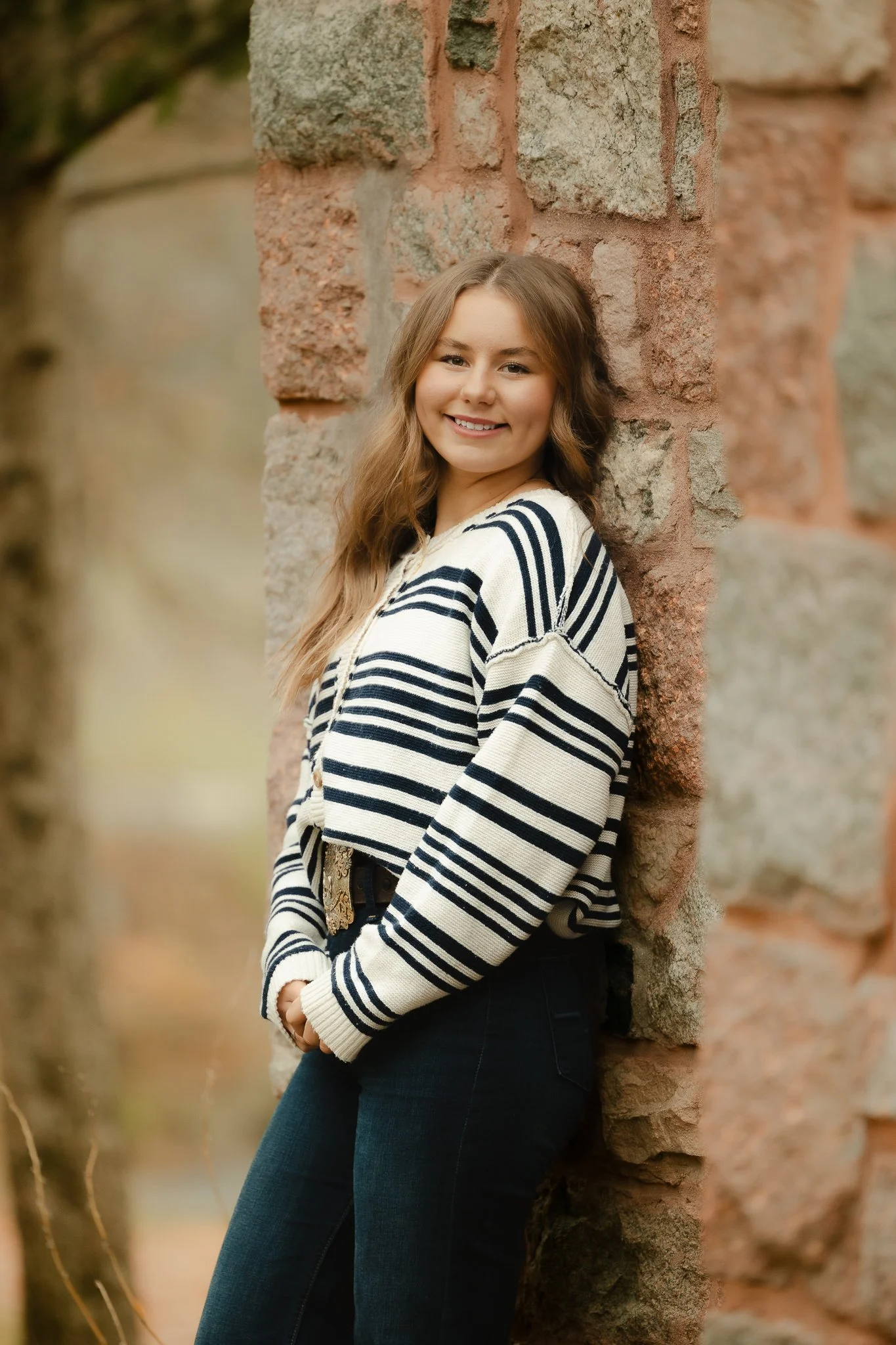 A young woman with long wavy hair smiling and posing against a stone wall outdoors.
