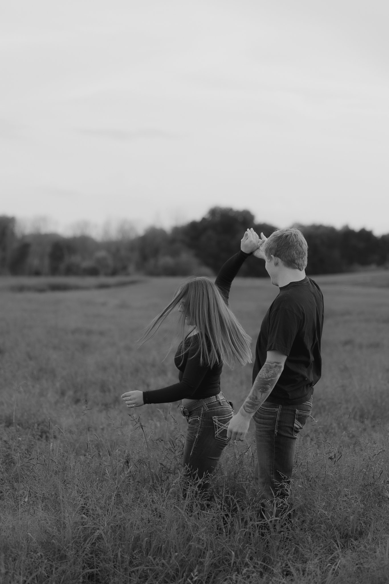 A black-and-white photograph of a young woman and man dancing or playing in a grassy field outdoors, with trees in the background