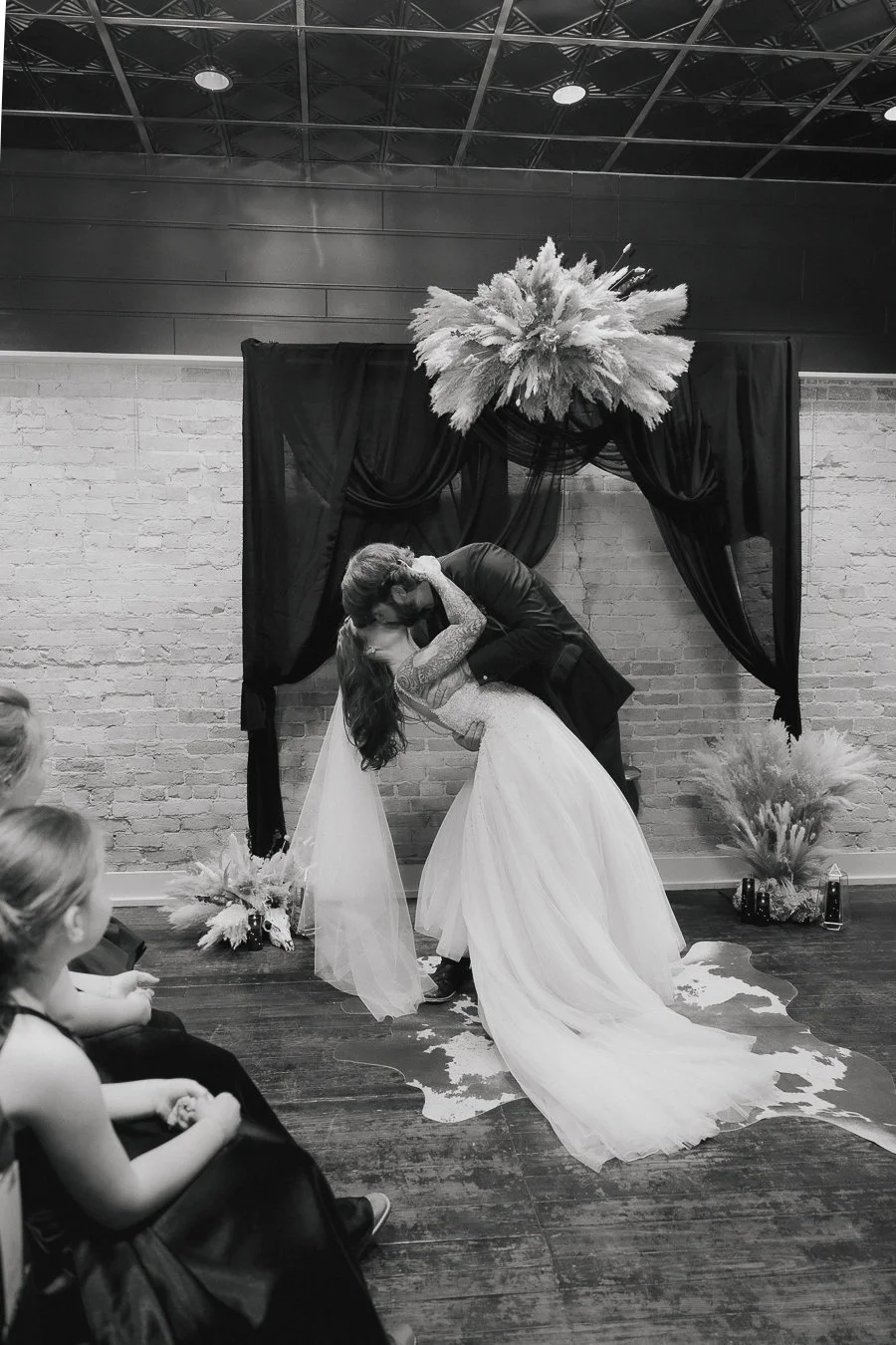 A bride and groom kissing during their wedding ceremony, with the groom dipping the bride, in front of a decorative backdrop with flowers, on a hardwood floor.