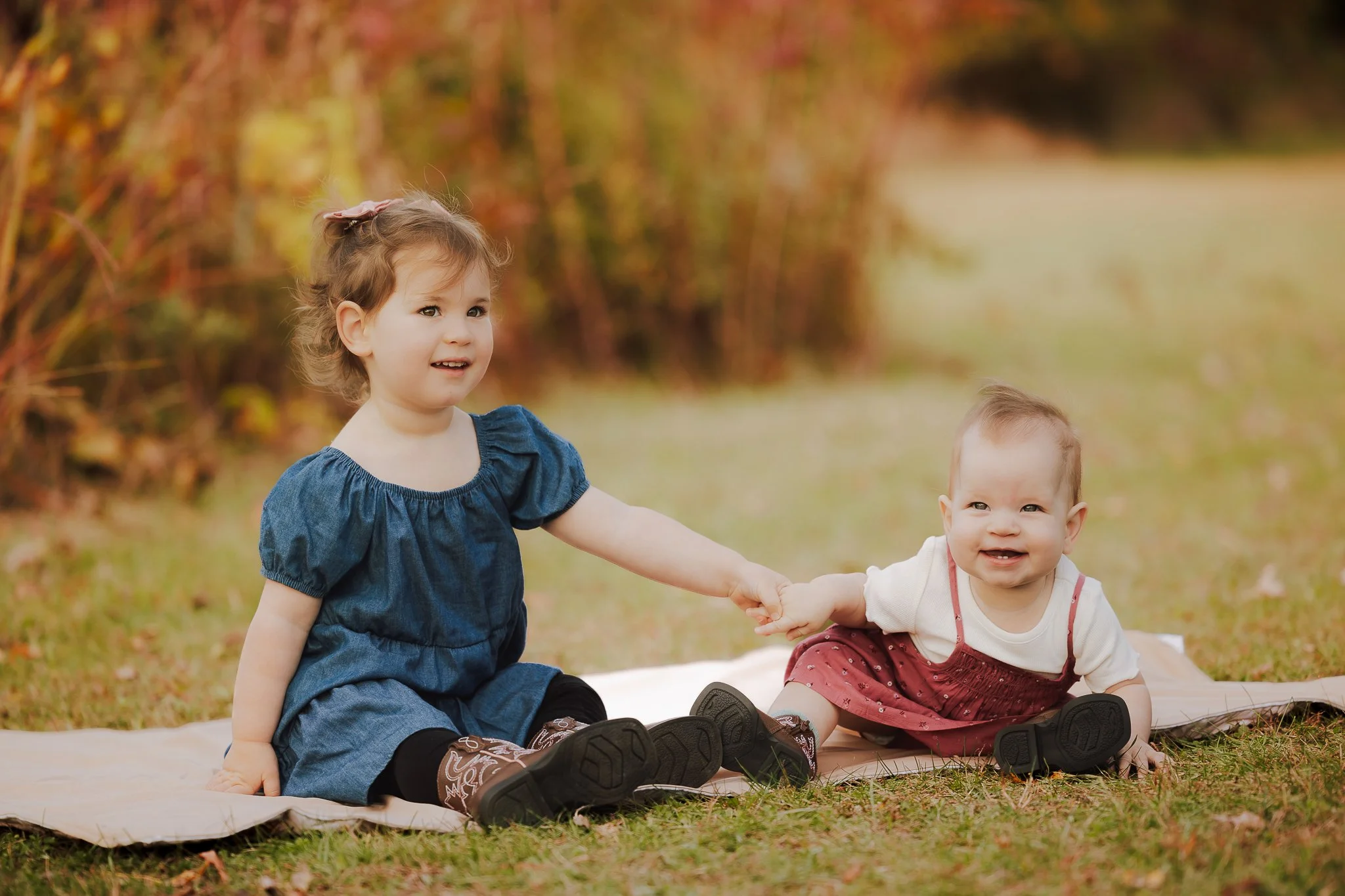 A young girl and a baby sitting on a blanket outdoors, holding hands and smiling, with autumn foliage in the background.