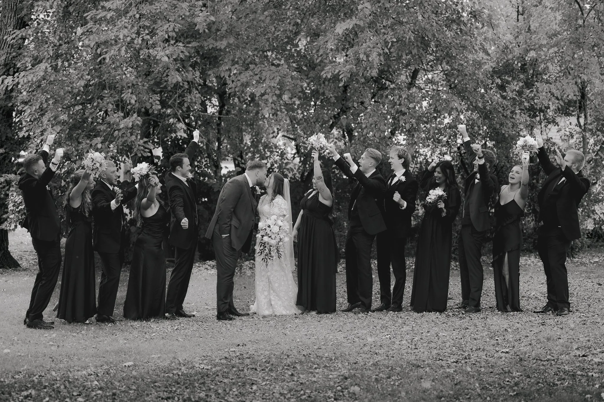 A black and white wedding photo of a couple kissing surrounded by friends, some holding bouquets, in a park with trees in the background.