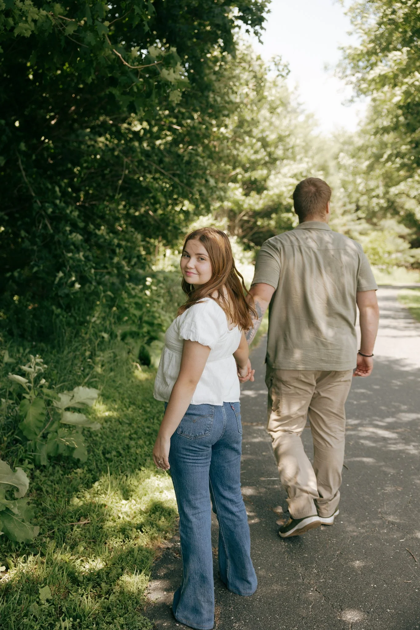A young woman with long auburn hair and a white top, smiling and holding hands with a man, while walking along a shaded outdoor path with green trees on either side.