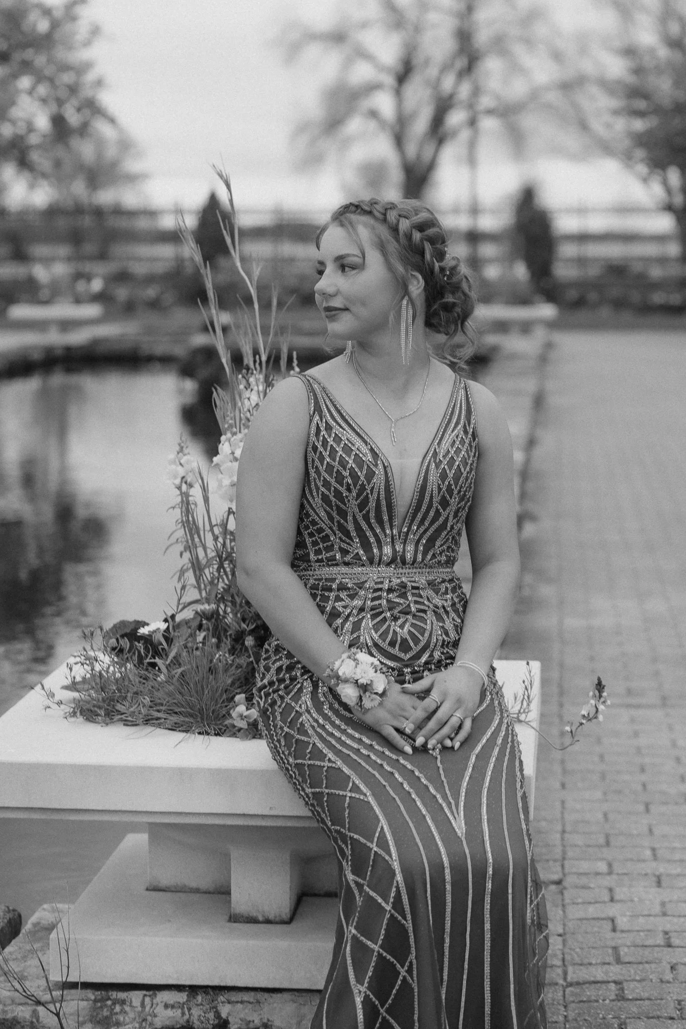 A woman in an ornate dress sitting on a concrete bench by a pond, with trees and blurred people in the background. She has earrings, a necklace, and flowers on her wrist, looking to the left.