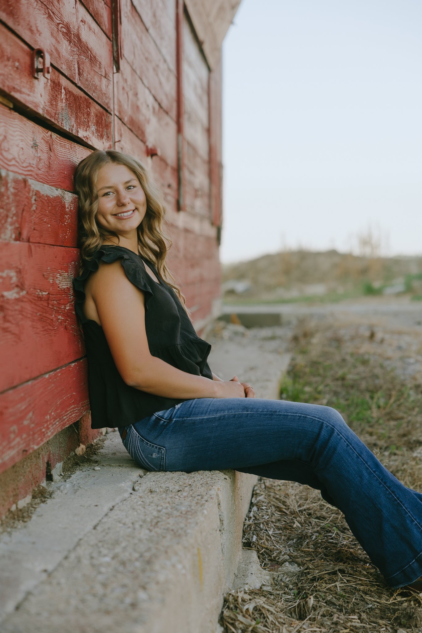 A young woman with wavy hair sitting on a concrete ledge beside a weathered red wooden building, smiling at the camera with a scenic outdoor background.