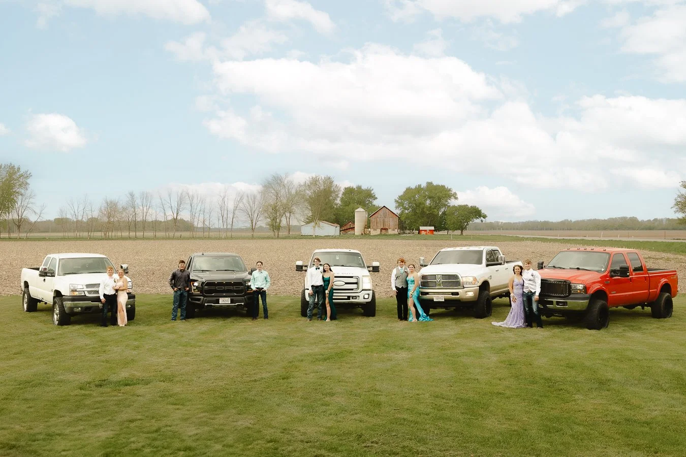 A group of people standing next to five pickup trucks in a grassy field with farmland and farm buildings in the background.