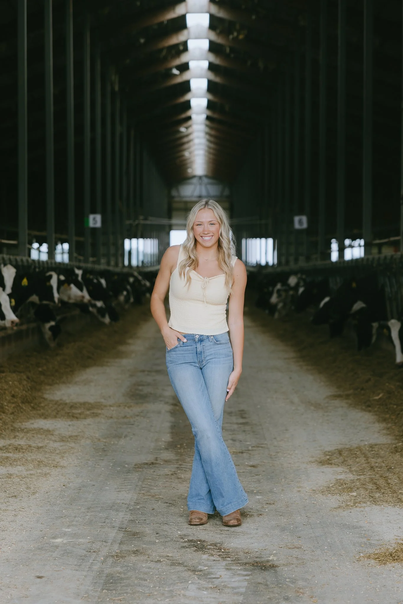 A young woman standing in the middle of a barn aisle with cows on either side, smiling and posing for the photo.