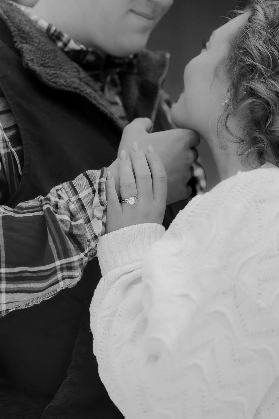 A couple holding hands close to each other, with a woman wearing an engagement ring, on a black and white photo.