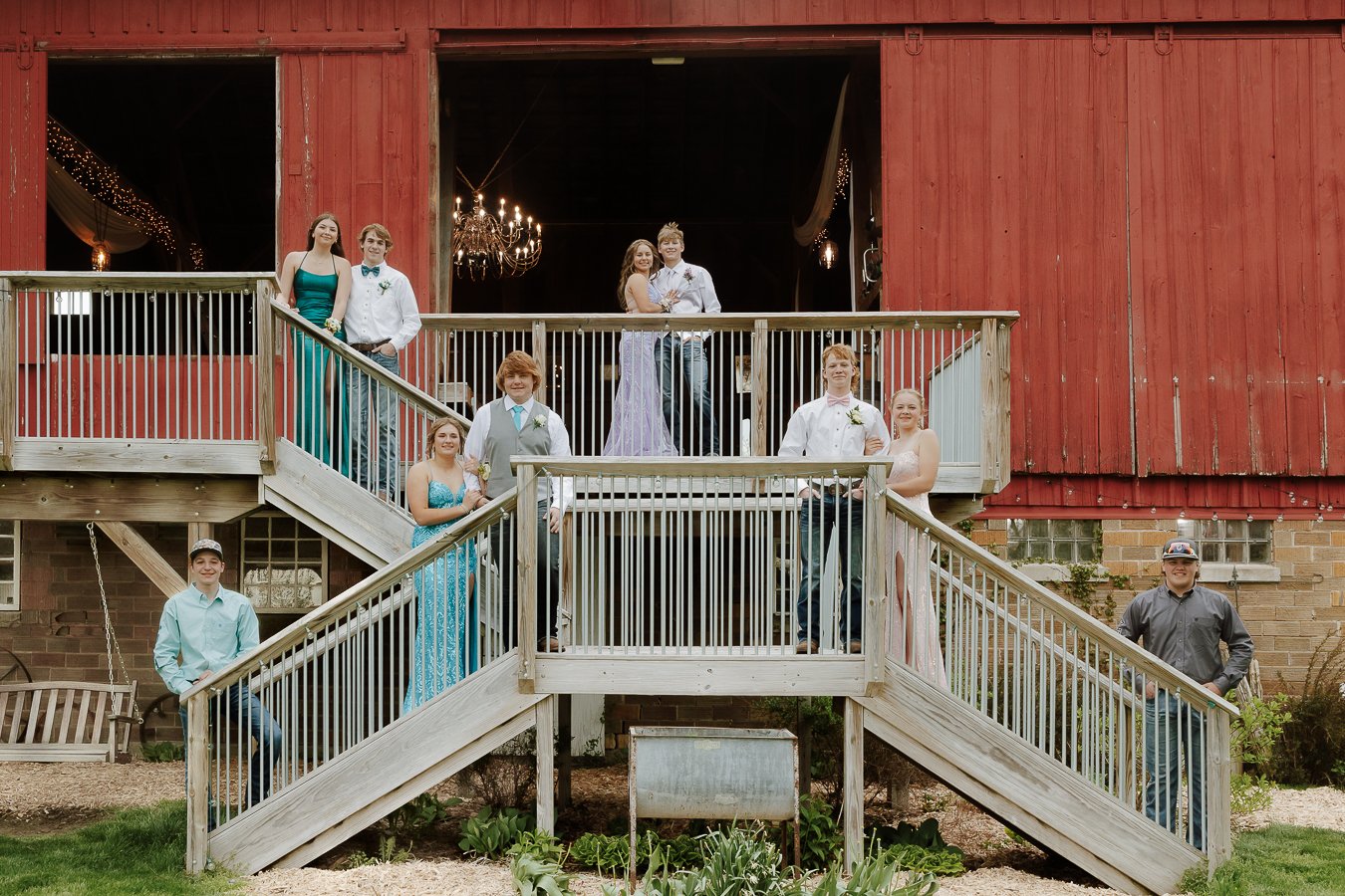 Group of teenagers dressed in formal attire standing on a wooden staircase outside a rustic red barn with a large open door and chandelier inside.