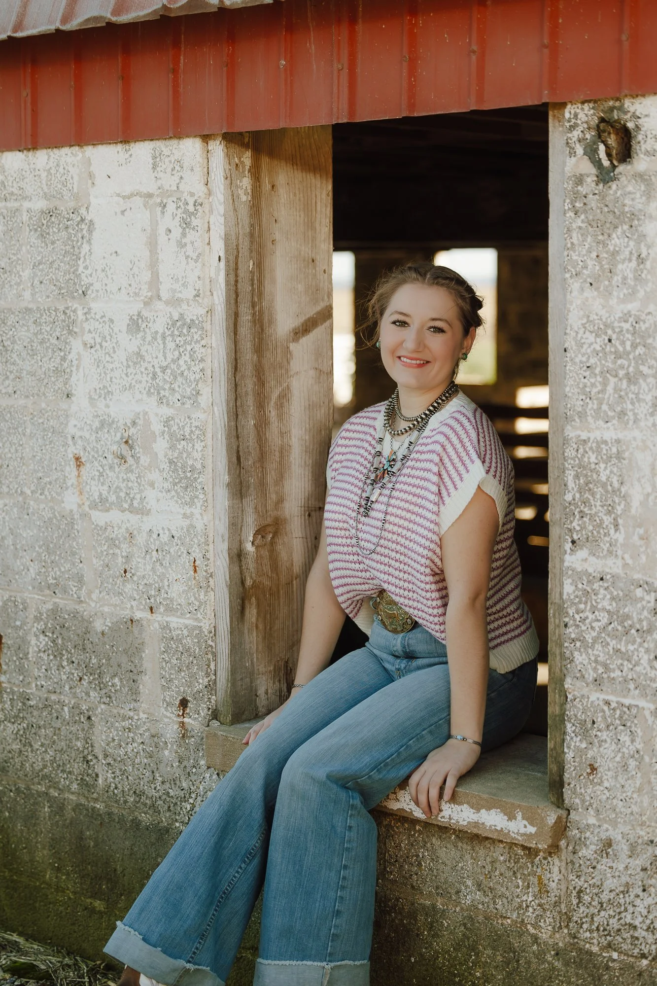 A young woman with braided hair sits at a rustic barn window opening, smiling at the camera. She wears a patterned knit top, light blue jeans, and layered necklaces, with a large belt buckle visible.