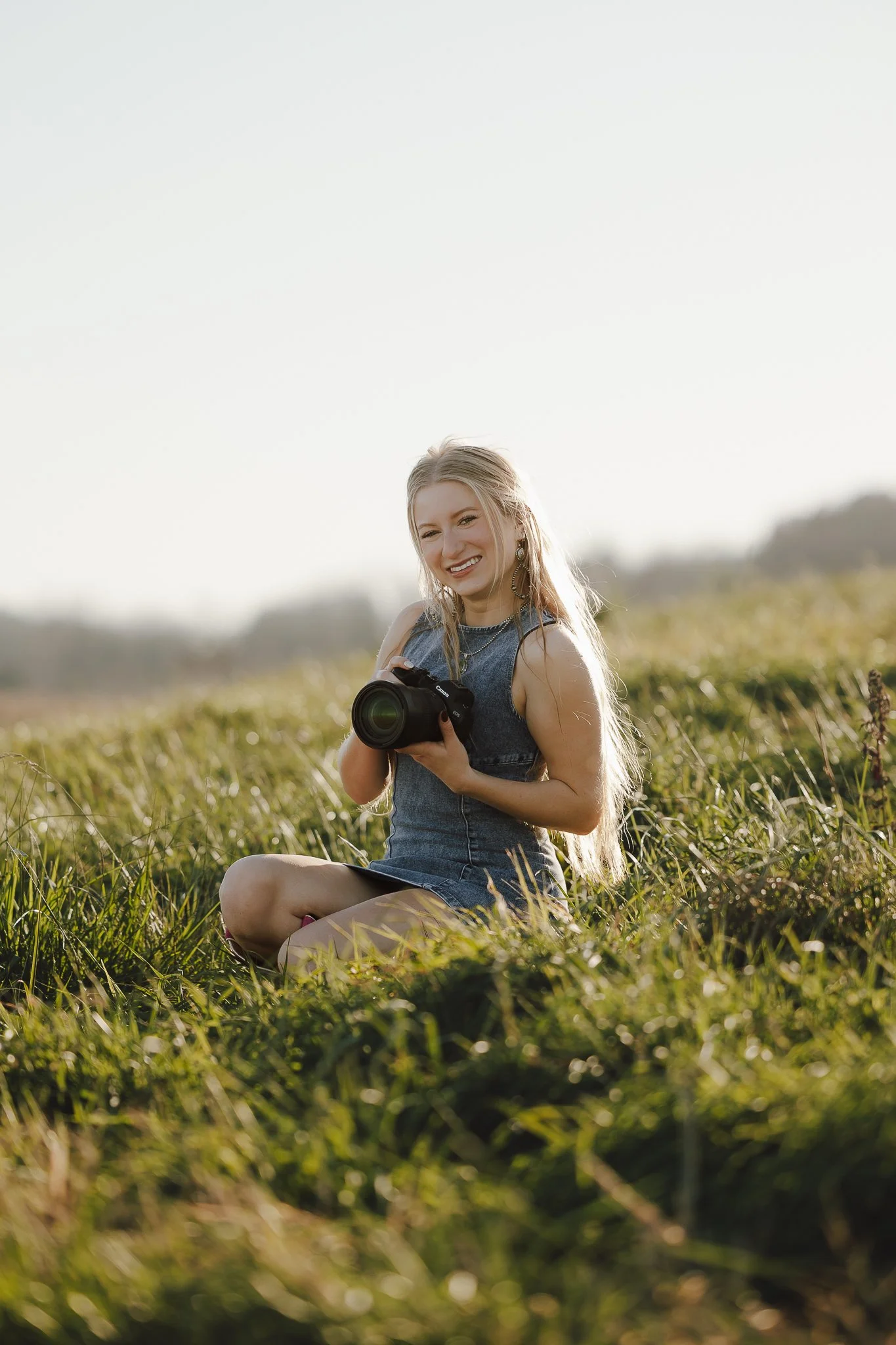 A young woman sitting in a grassy field, holding a camera and smiling at the camera.