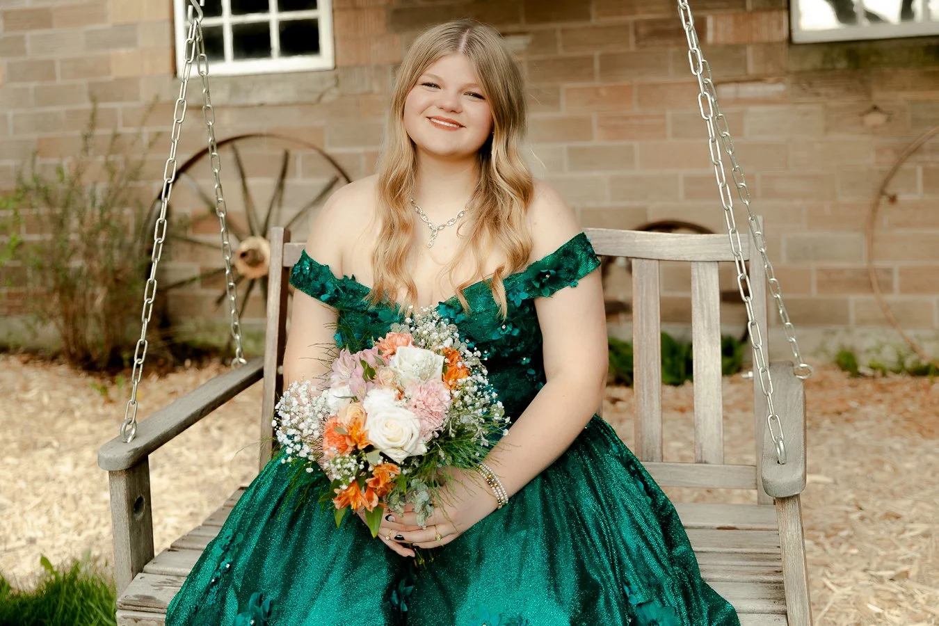 Young woman in a dark green, off-the-shoulder dress sitting on a wooden swingset, holding a bouquet of flowers, smiling outdoors with brick wall and wagon wheel in background.
