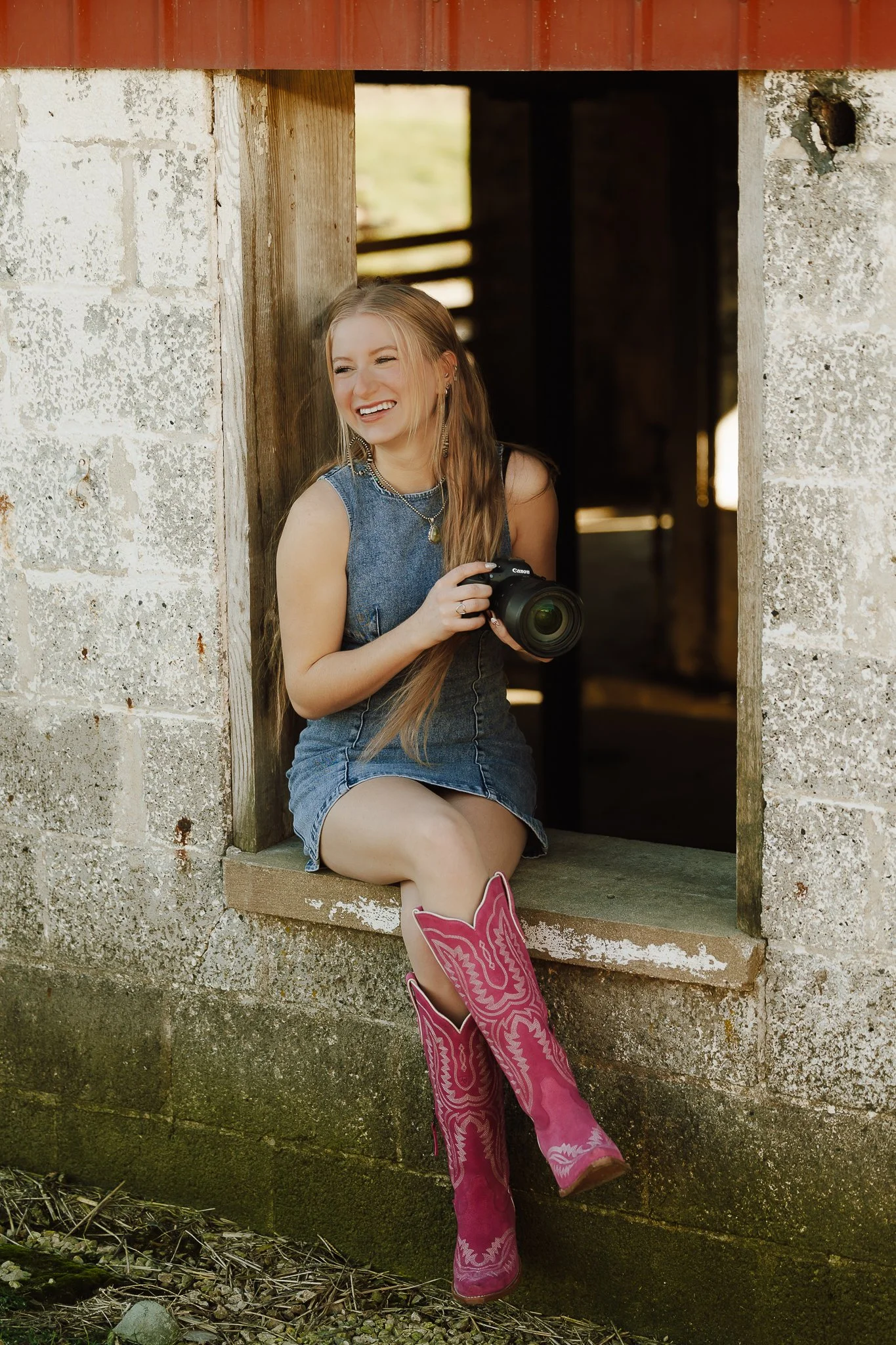 A young woman with long hair, wearing a denim dress and pink cowboy boots, is sitting on a ledge next to an open barn window, holding a camera and smiling.