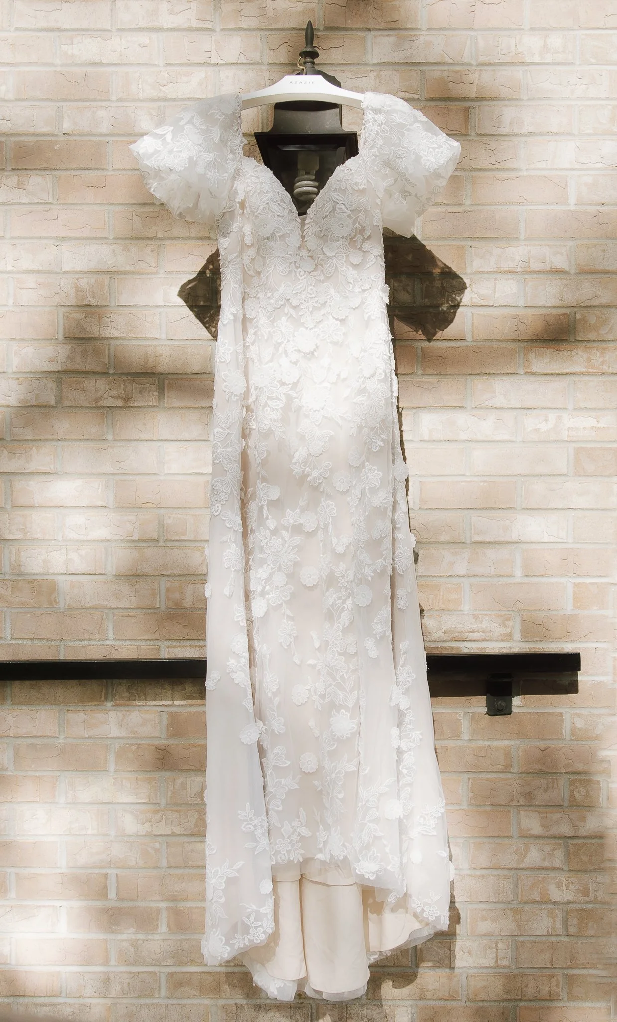 White lace wedding dress hanging on a hanger against a brick wall.