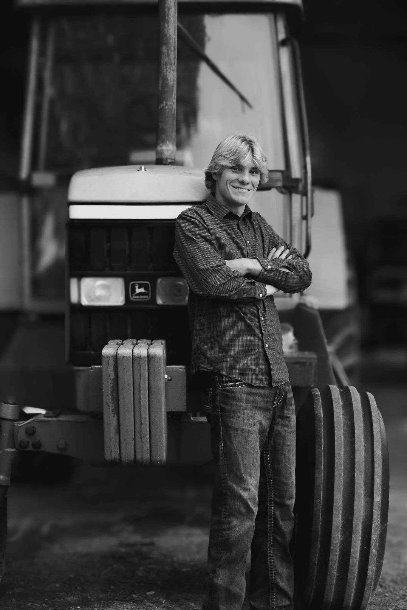 A young man with blonde hair, wearing a checkered shirt and jeans, standing in front of a tractor with arms crossed, smiling.