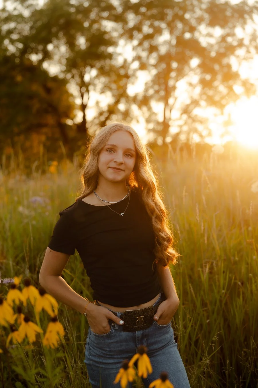 Young woman standing in a field of yellow flowers with tall grass, during sunset, wearing a black shirt and jeans, smiling softly.