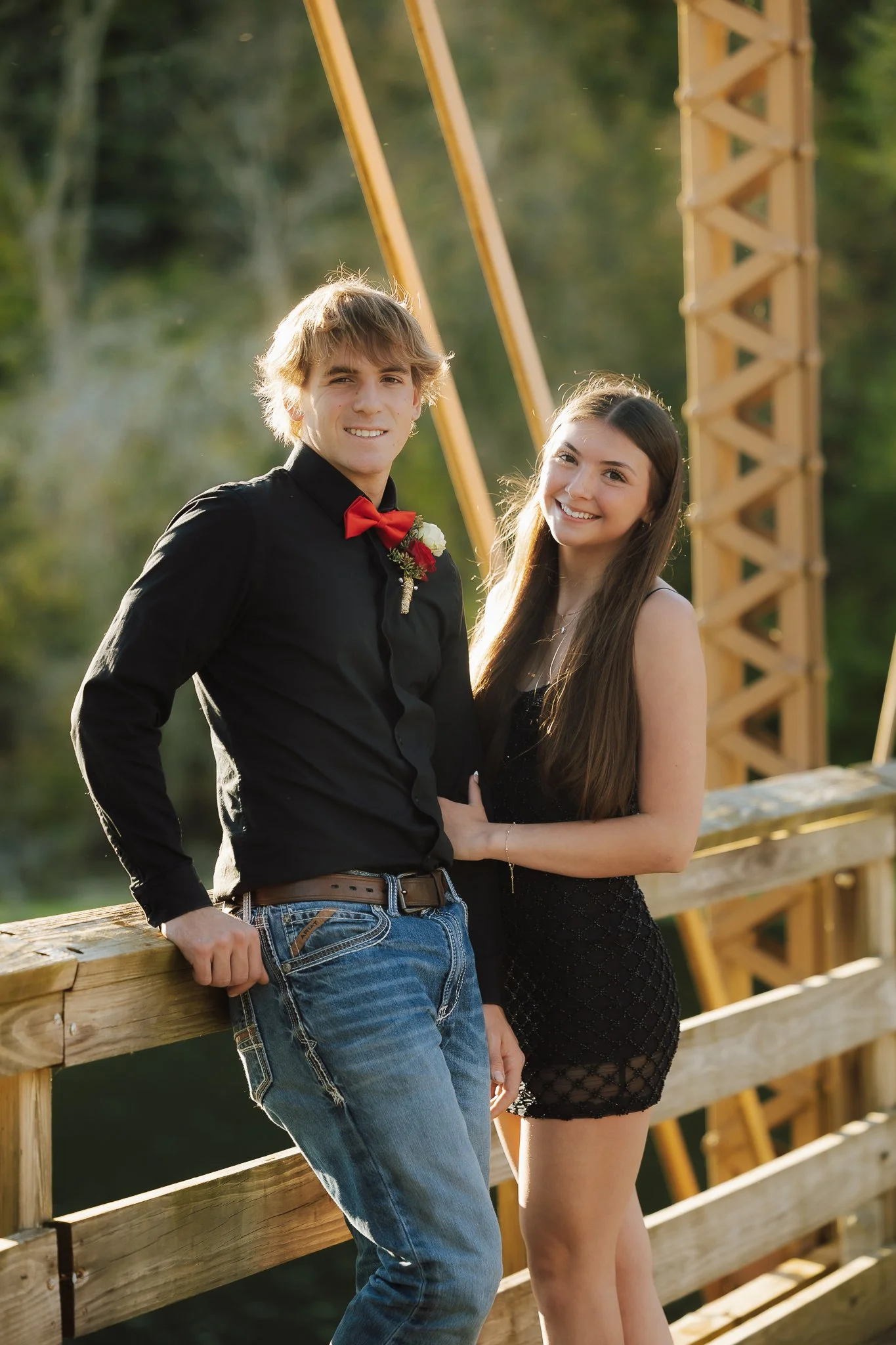 A young man and woman standing together on a wooden bridge outdoors, smiling at the camera, with trees and a yellow bridge tower in the background.