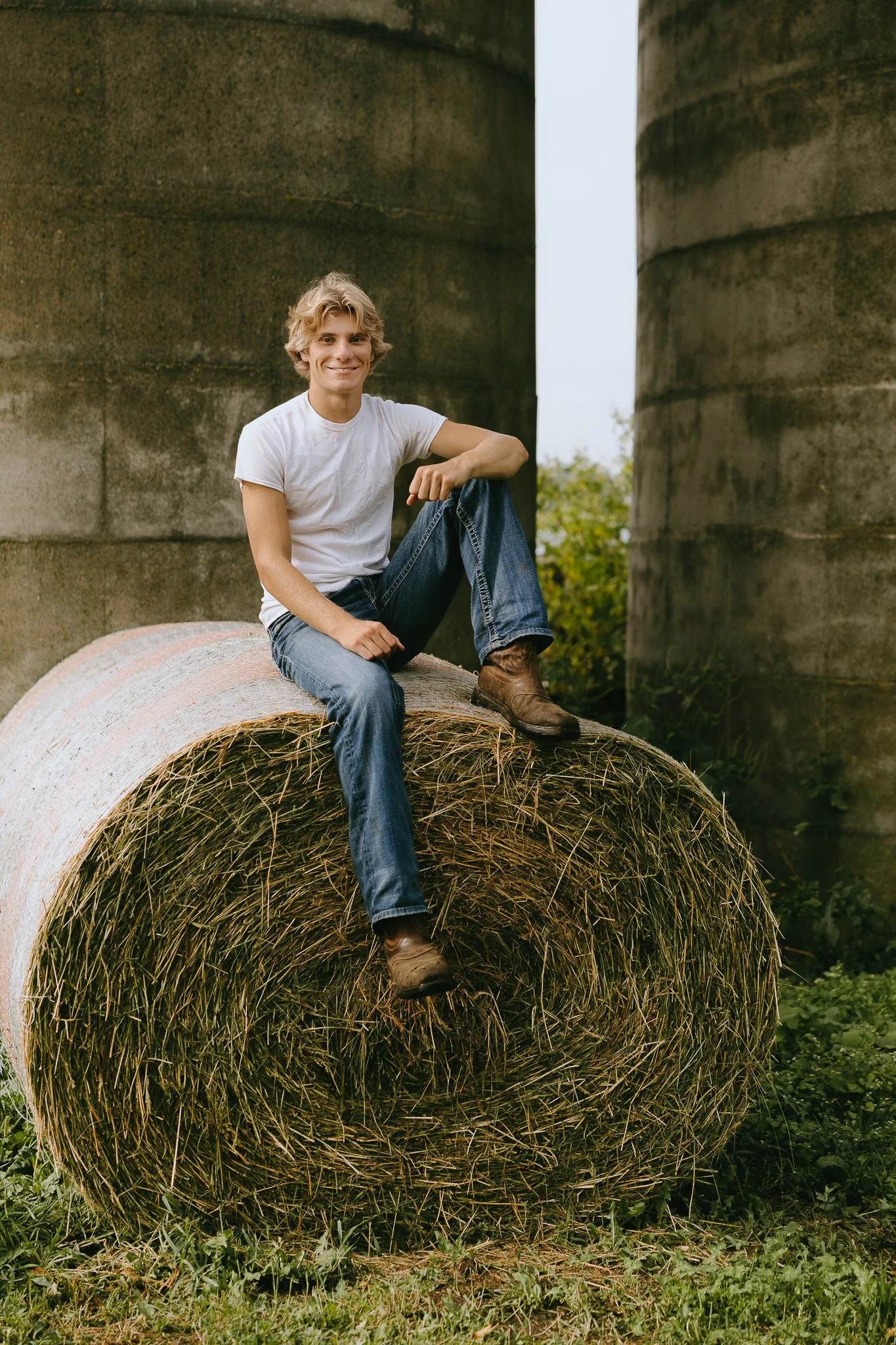 A young man in a white t-shirt, blue jeans, and brown boots sitting on a large round hay bale outdoors, with stone silos in the background.