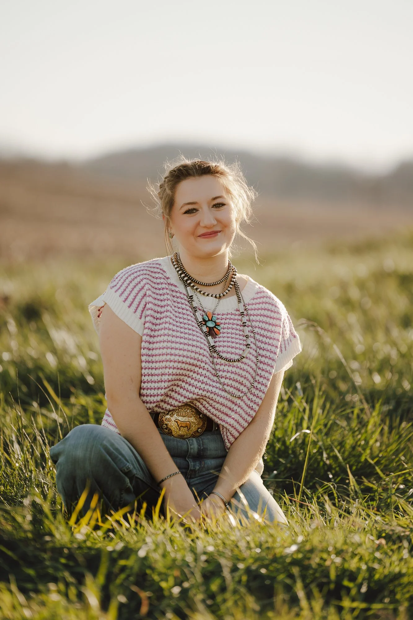 A young woman sitting on grass outdoors, smiling, wearing a pink and white striped top, jeans, layered necklaces, and a belt with a large belt buckle, with a background of blurred hills and a clear sky.