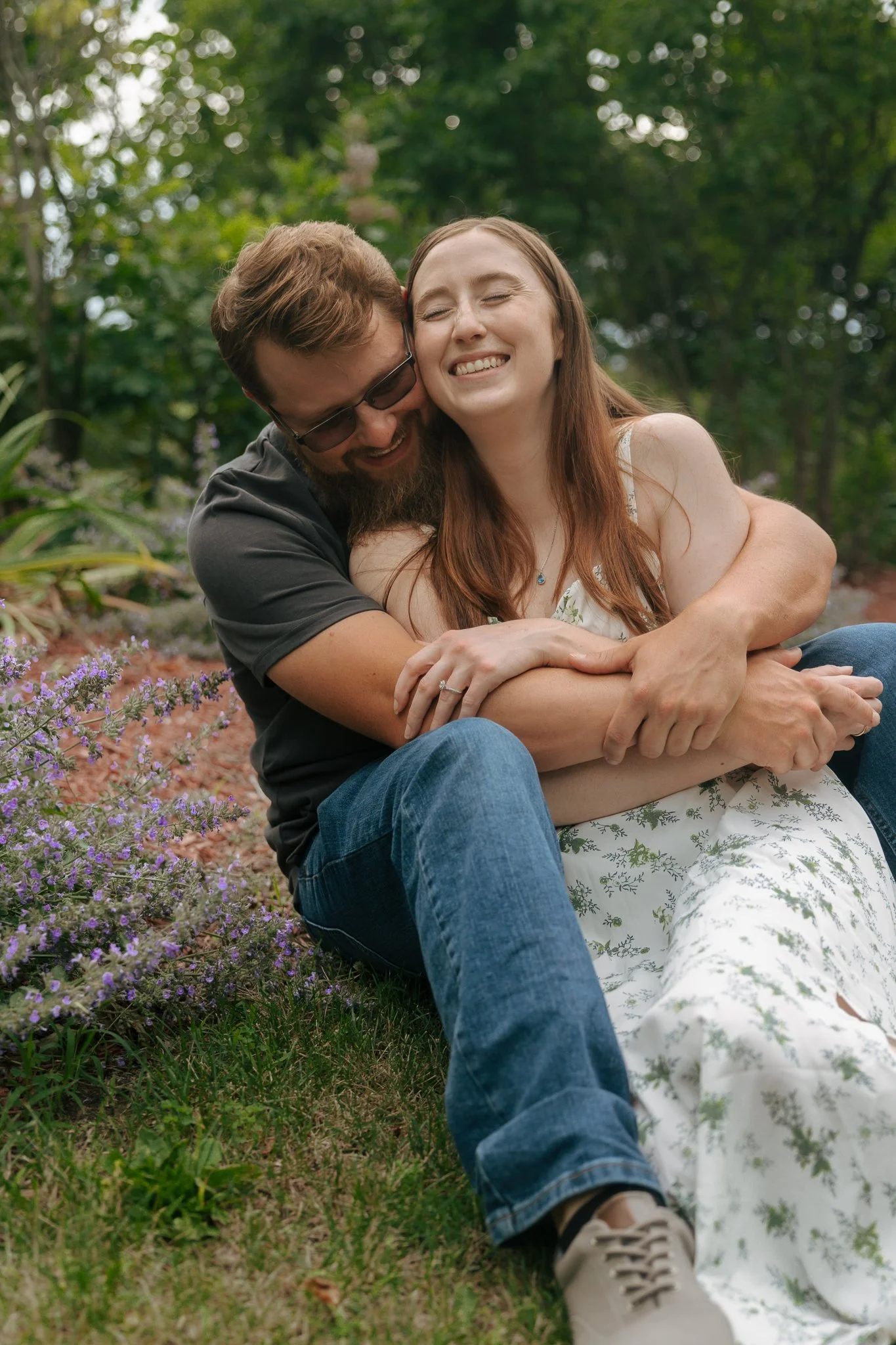 A couple sitting on grass in a garden, hugging and smiling. The woman has long red hair, and the man has short hair, a beard, and sunglasses. Purple flowers are nearby, and green trees are in the background.
