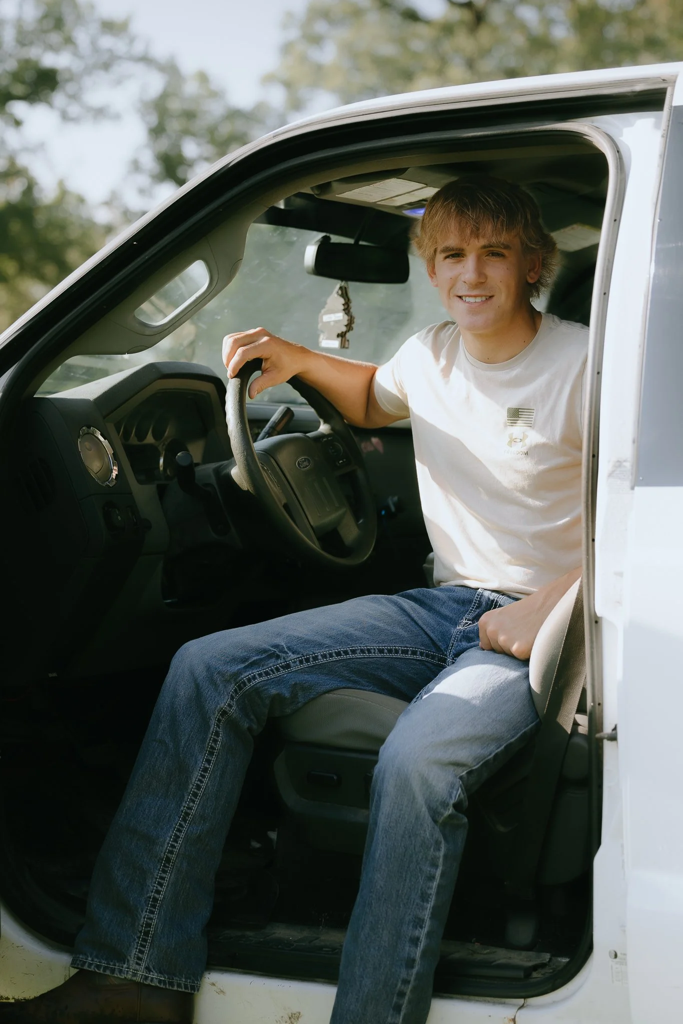 Young man sitting in the driver's seat of a white vehicle, smiling, wearing a white t-shirt and blue jeans, with the window rolled down on a sunny day.