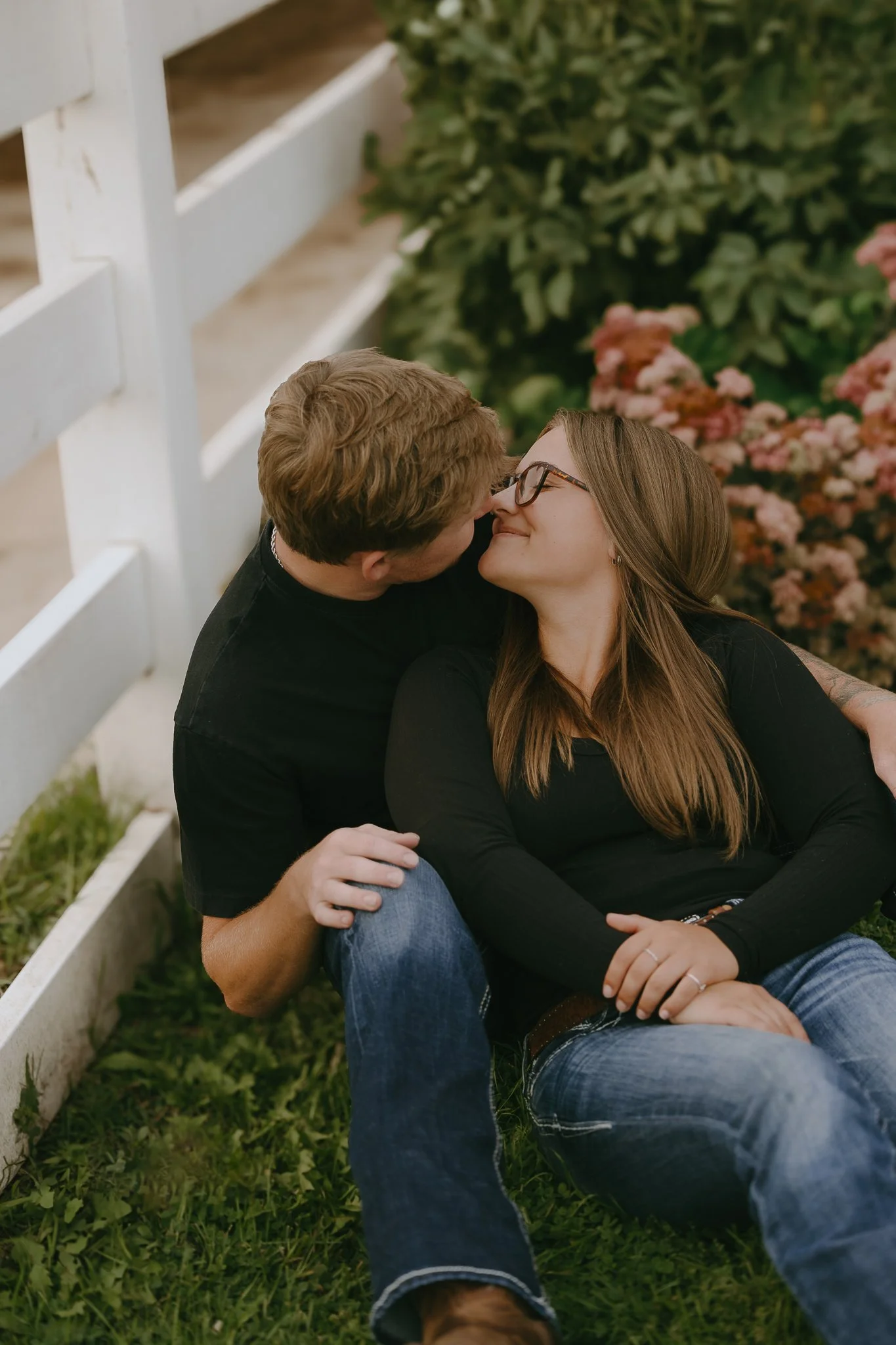 A couple sitting on grass, leaning against a white fence, sharing a kiss. The woman has long hair, glasses, and is wearing a black top and blue jeans. The man has short hair and is wearing a black shirt. The background includes some flowering bushes.