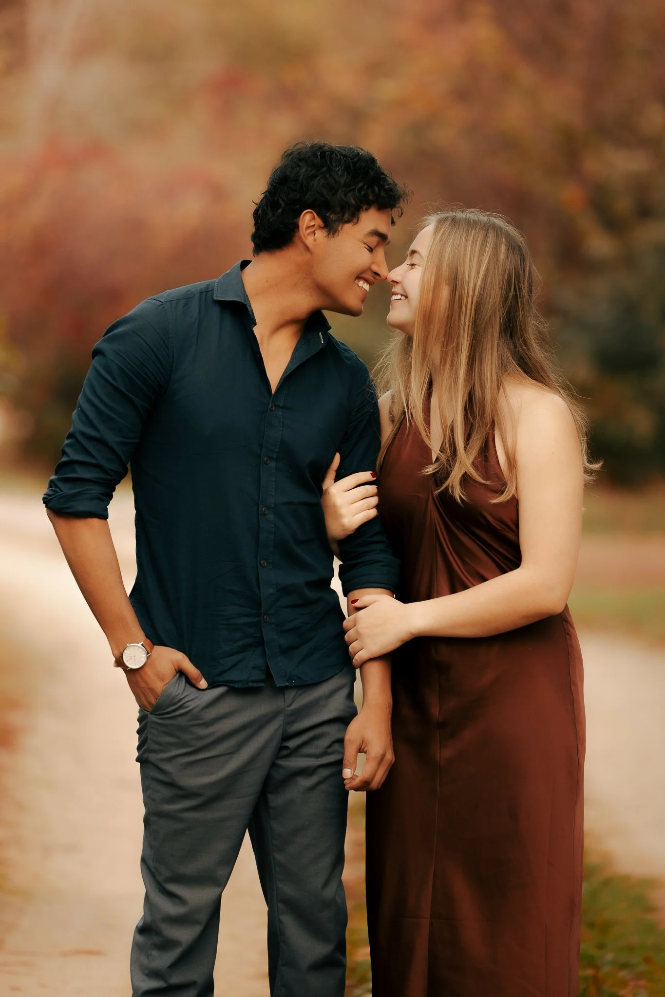 A young couple smiling closely, about to kiss, standing outdoors surrounded by fall foliage.