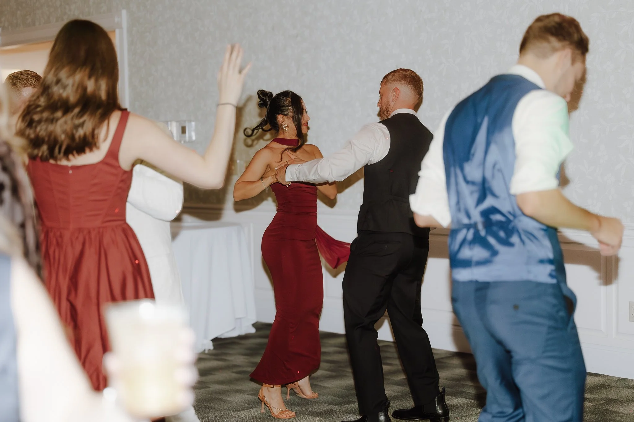 People dancing at a celebration, two women and two men prominent in the scene, dressed in formal attire, with one woman in a red gown and the other in a white dress, and the men in white shirts and suits.
