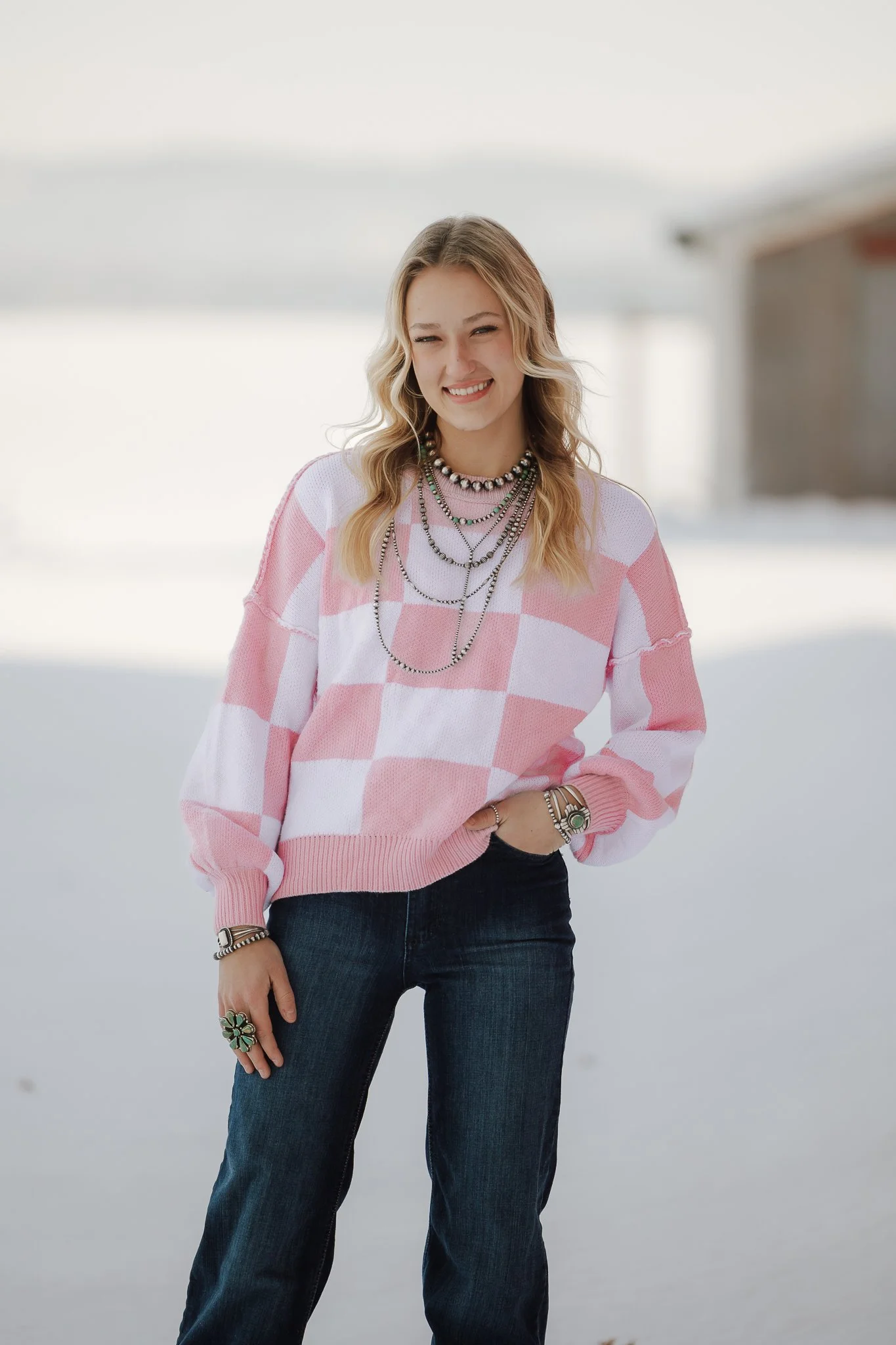A young woman with blonde hair is standing outdoors in a bright, snowy setting, smiling with her eyes partly closed, wearing a pink and white checkered sweater, several layered necklaces, and dark jeans.