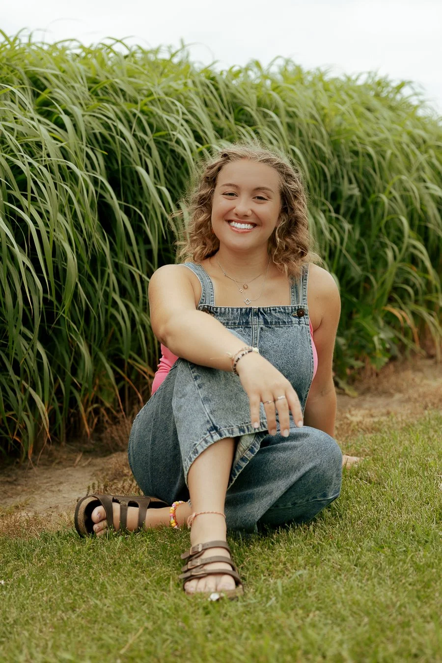 Young woman with curly hair smiling, sitting on grass, wearing denim overalls, pink top, and sandals, in front of green plants.