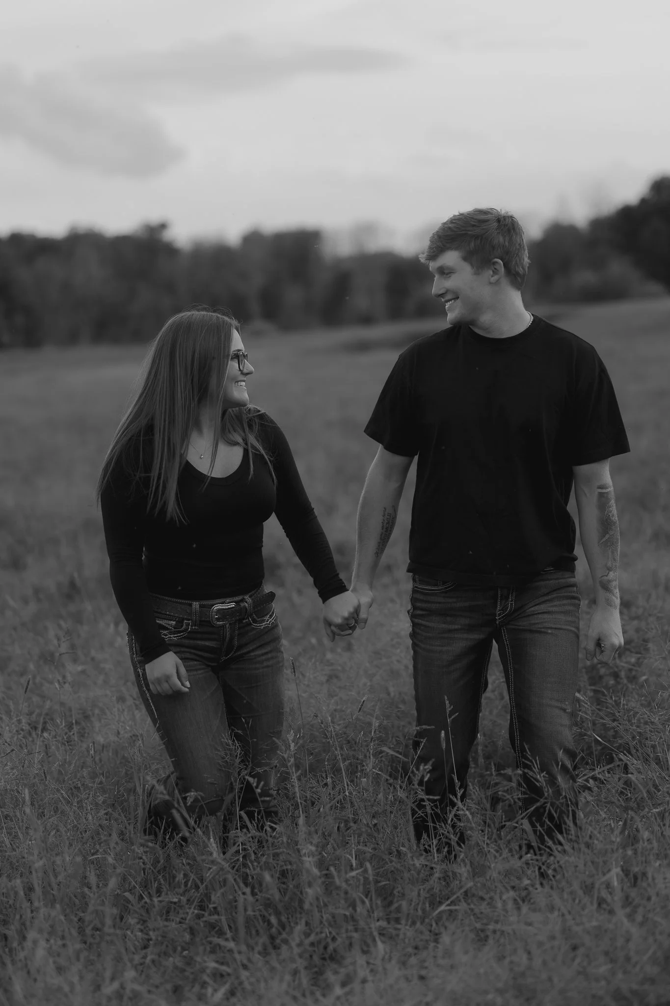 A black and white photograph of a smiling couple holding hands while walking through a grassy field, looking at each other.
