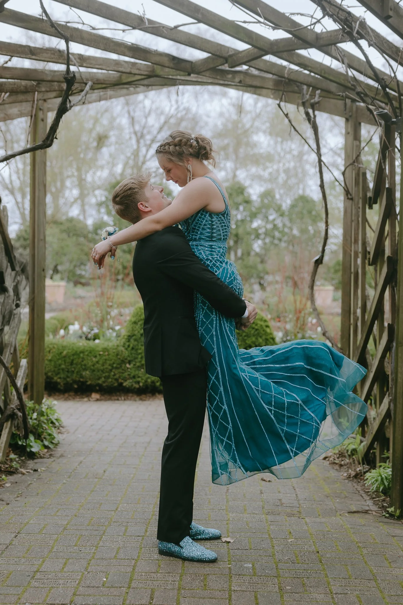 A young man in a black tuxedo lifting a young woman in a teal dress in an outdoor garden setting, both gazing at each other lovingly.