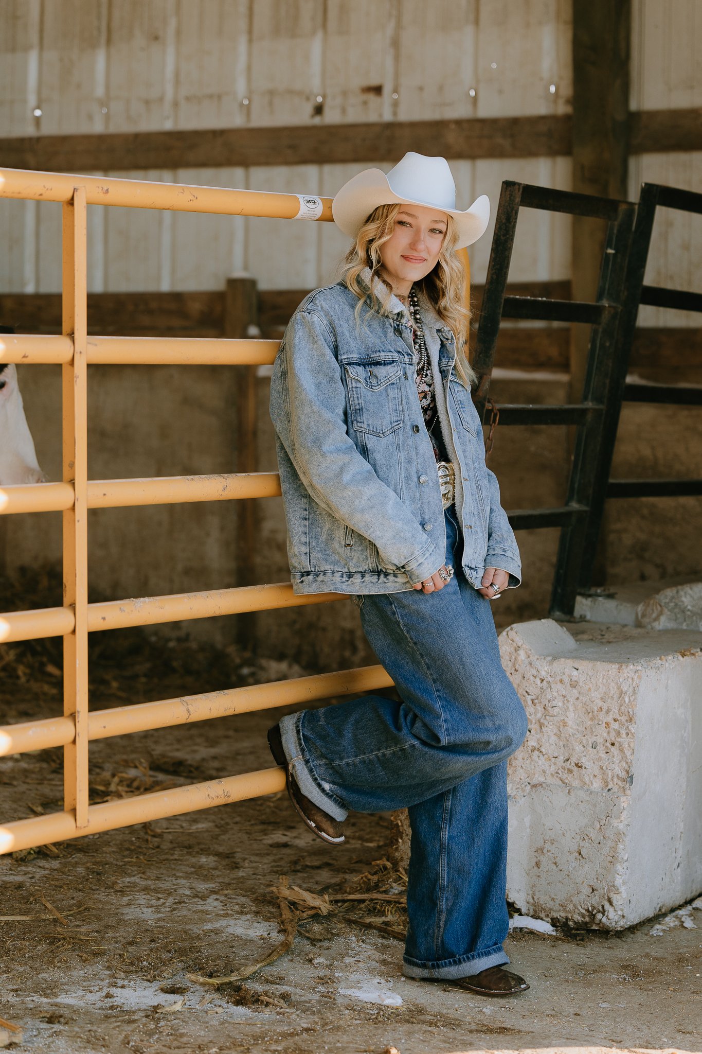 Young woman in cowboy hat, denim jacket, and jeans leaning against a yellow metal fence inside a barn.