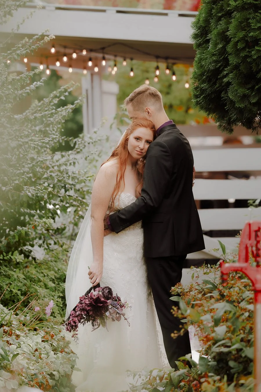A bride and groom embracing outdoors, surrounded by lush greenery, with wedding decorations and string lights hanging above.