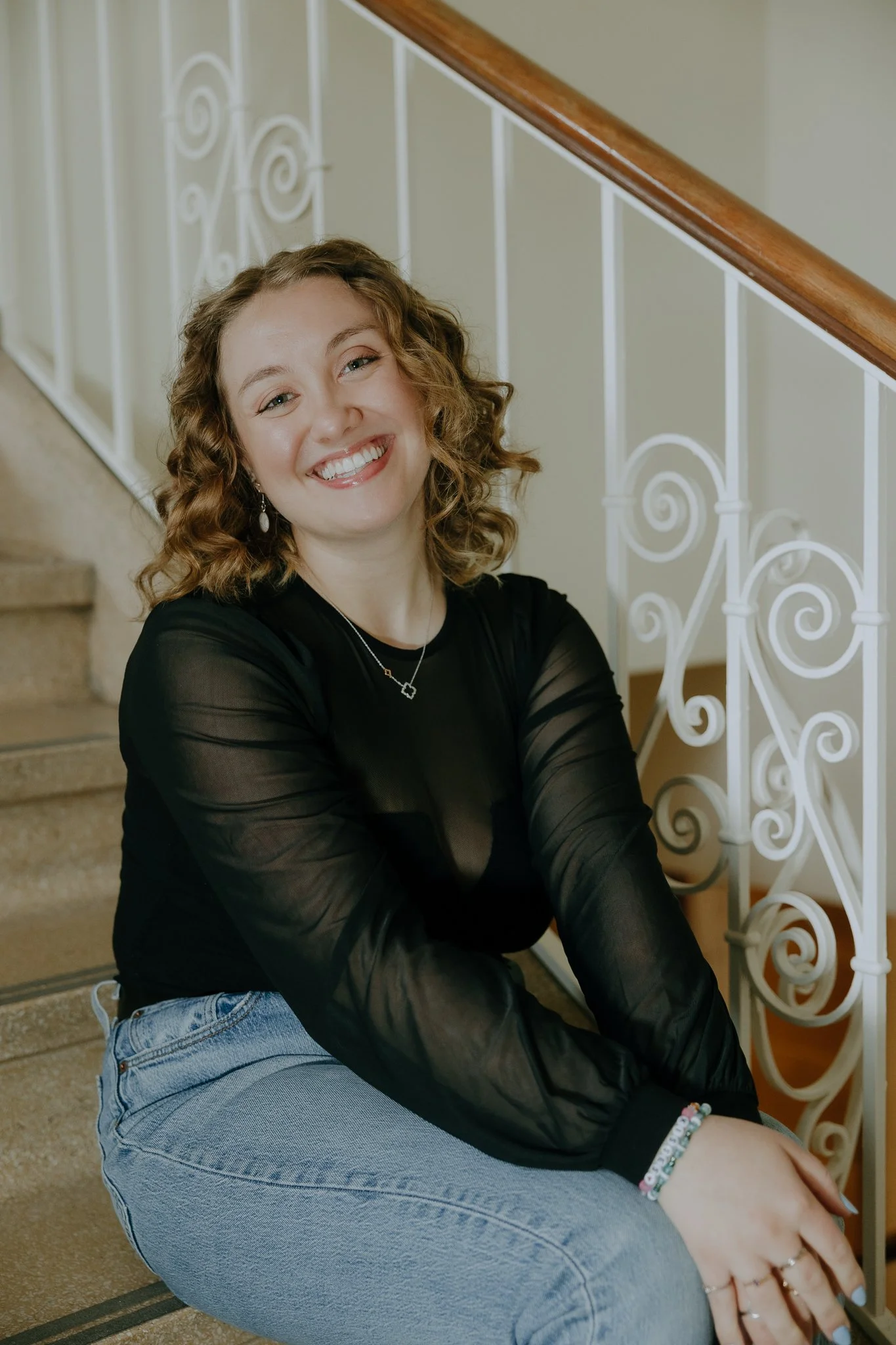 A young woman with curly blonde hair sitting on stairs, smiling at the camera, wearing a black sheer long-sleeve top, light blue jeans, earrings, and bracelets.