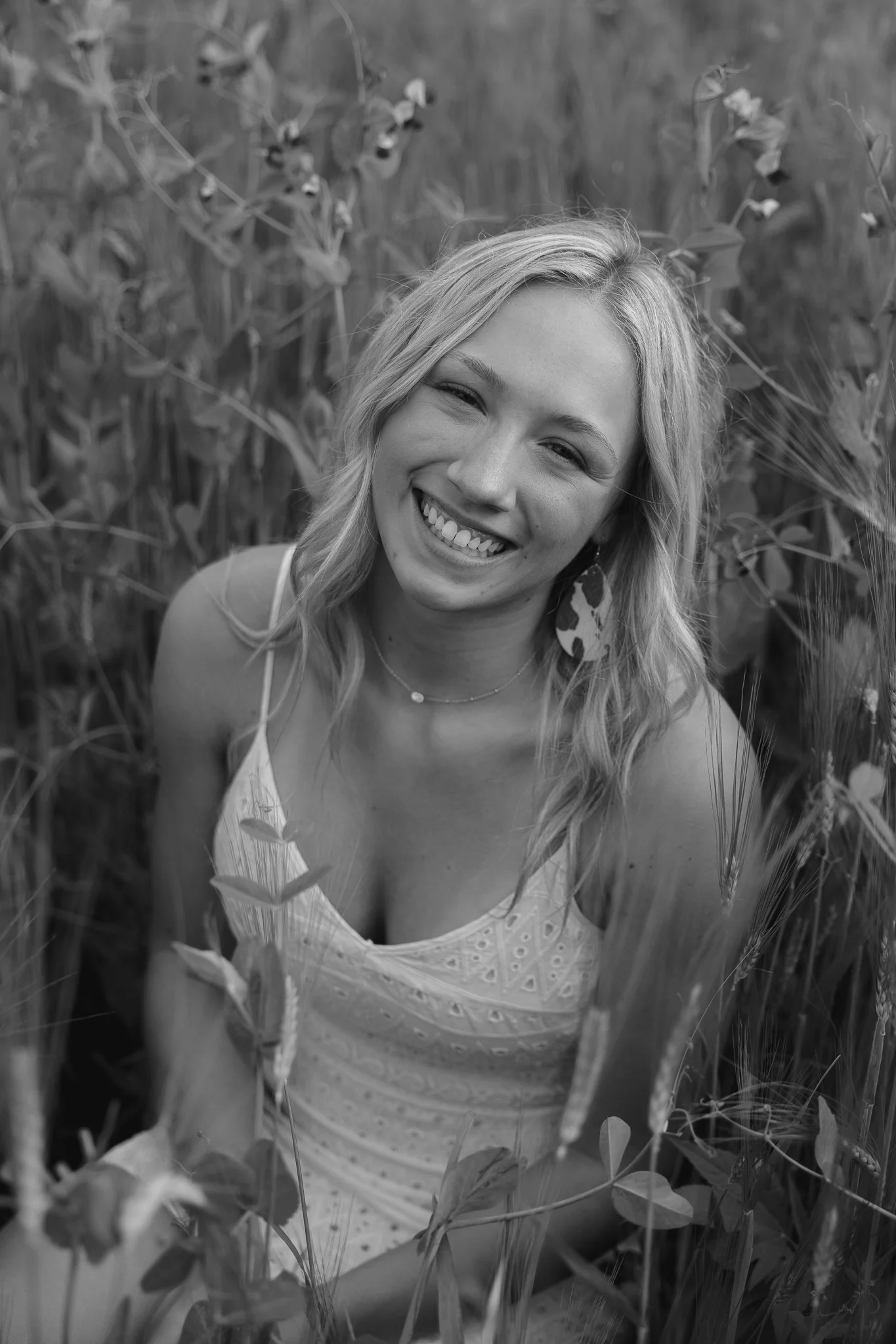 A young woman with blonde hair smiling in a field of tall grass and plants, wearing a sleeveless dress and a necklace.