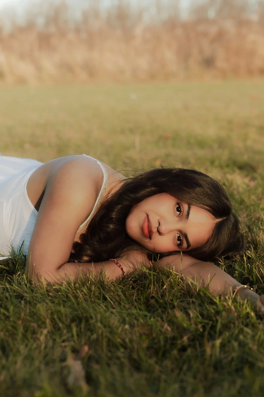 Young woman with long dark hair lying on grassy field, resting her head on her arm, smiling softly with a natural outdoor background.
