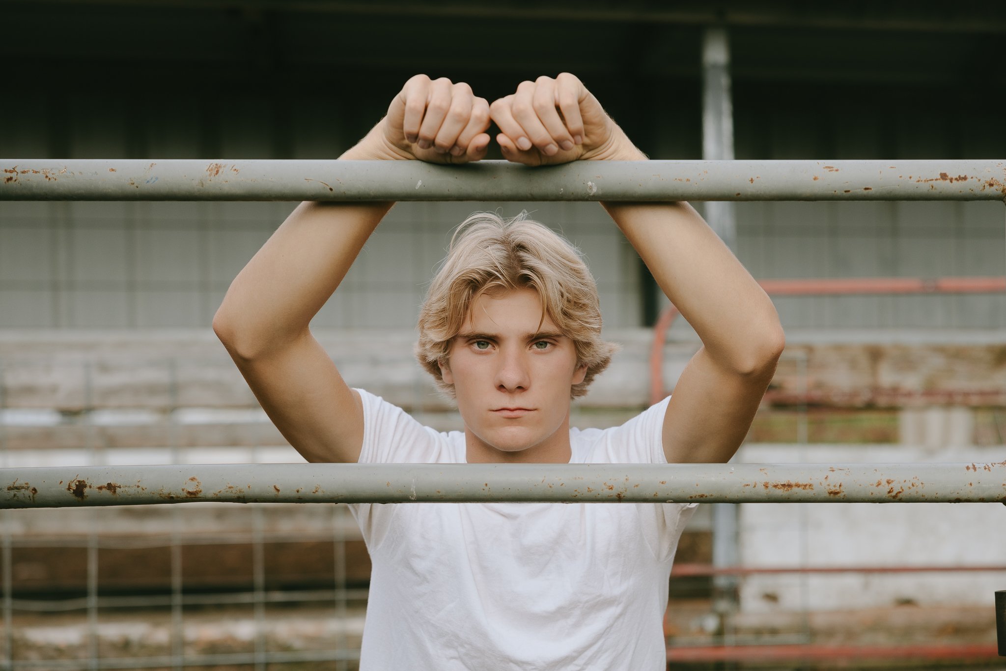 Young man with blonde hair, standing outdoors behind metal bars, looking directly at the camera with a serious expression.